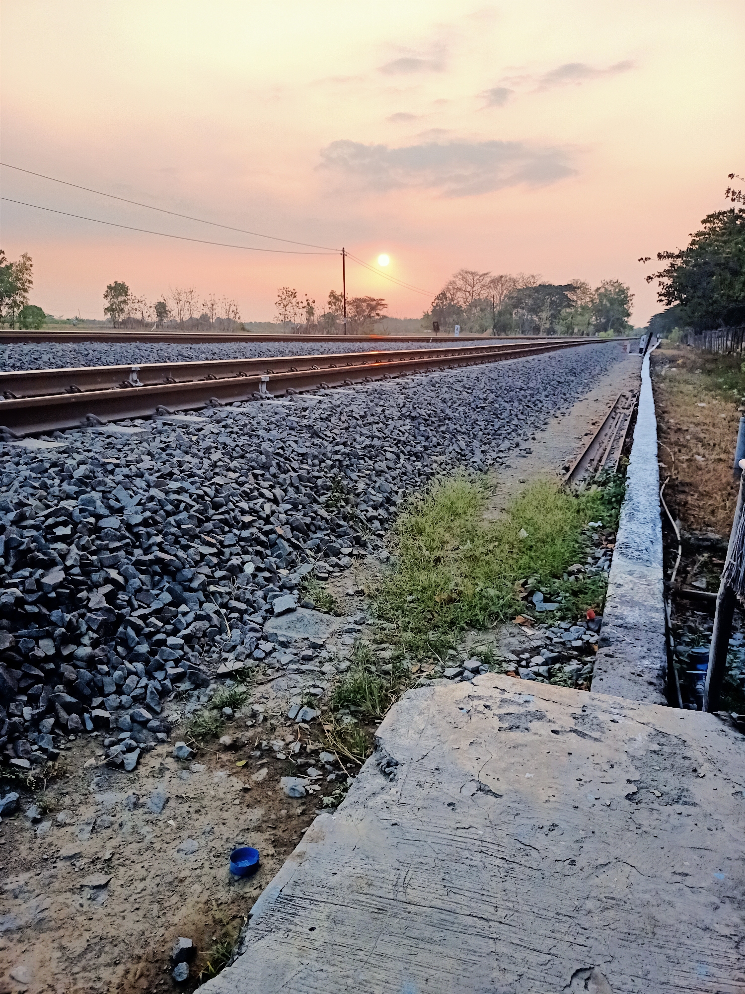 The image depicts a railway track in Banjarjo, Indonesia, during a sunset. The sky is filled with soft orange and pink hues, with a few scattered clouds. The sun is visible as a bright orb near the horizon, casting a warm glow over the landscape. The railway tracks stretch into the distance, flanked by ballast stones and sparse vegetation. In the foreground, a concrete structure partially occupies the lower right portion of the frame. A blue plastic cap is visible on the ground near the tracks. The environment appears rural, with trees dotting the background. There are no people or animals visible in the image.