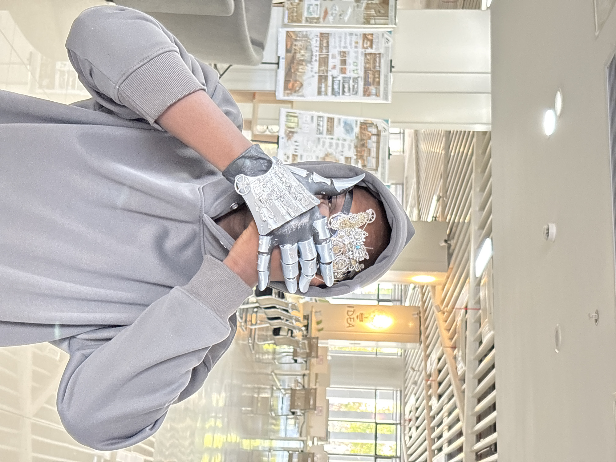 A person in a grey hooded sweatshirt is covering their face with their hands. One hand is covered by a silver metallic glove with intricate, mechanical designs. The location appears to be a modern interior space, possibly a library or academic building, with bookshelves, large windows, and displayed architectural drawings. The city of Jiangjin, China, is not explicitly identifiable from this image.
