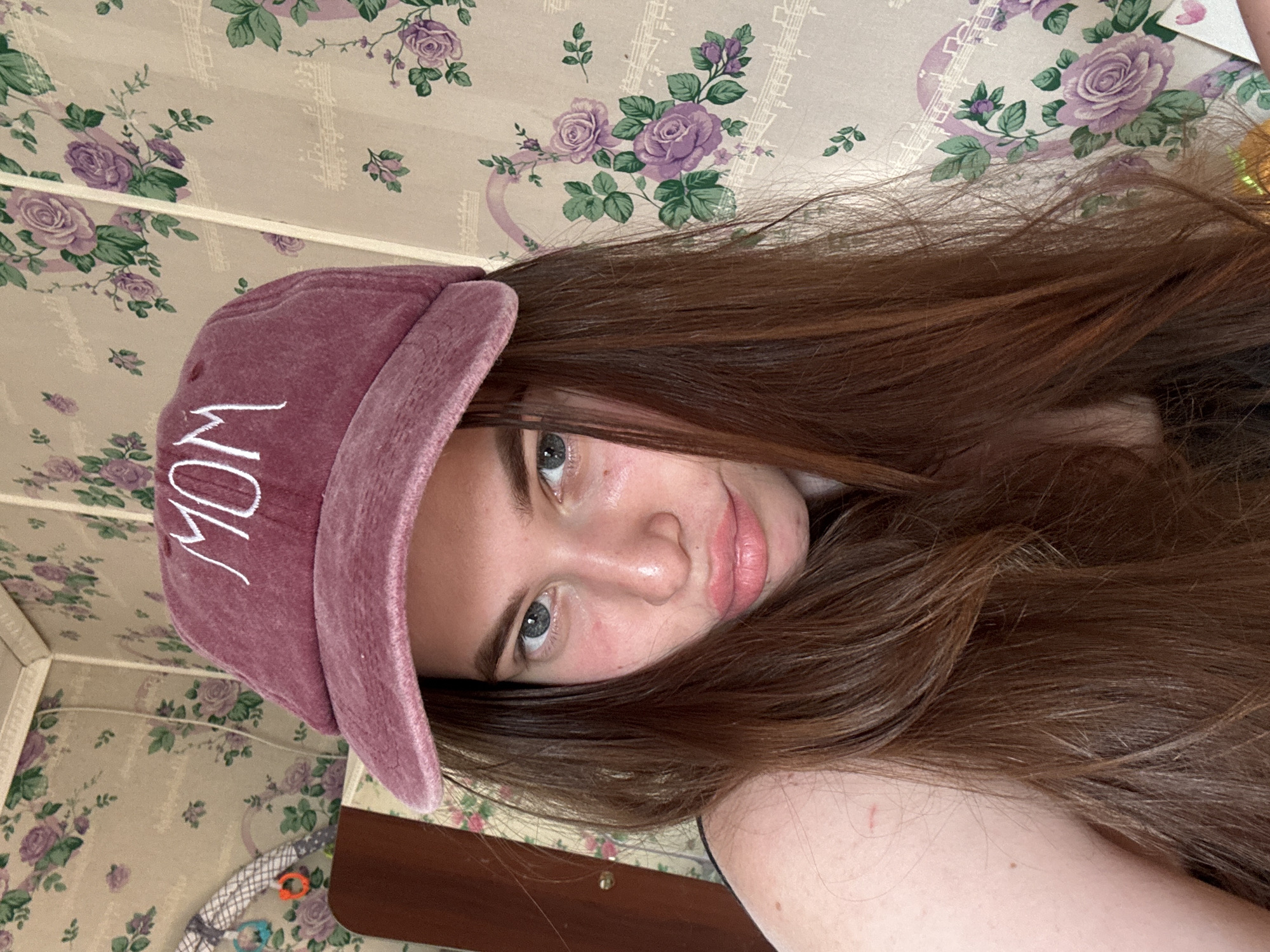 A woman with long, brown hair and grey eyes is wearing a pink baseball cap with the word "MOM" embroidered in white. The woman's skin tone appears fair with some redness on her face. She is lying on a bed with a floral-patterned comforter with lavender roses. The scene appears to be indoors, the specific location of Мещёры, Russia is indeterminable.