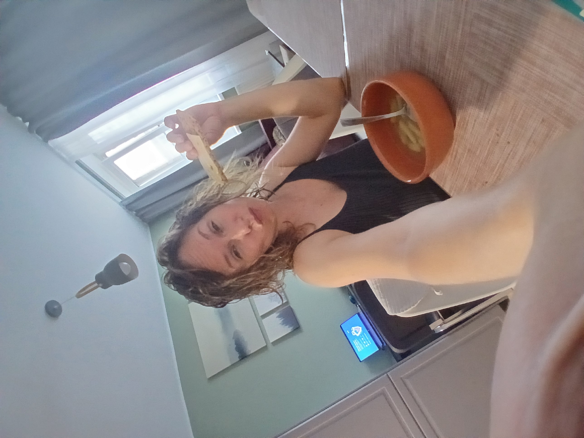 The image depicts a woman sitting at a table, holding a piece of bread up to her mouth and looking at the camera. She is wearing a dark, sleeveless top. In front of her, on the wooden table, is an orange bowl containing food and a spoon. The background shows a room with a laptop on a desk, a window with curtains, a light fixture on the ceiling, and artwork on the wall. Without specific visual cues or landmarks, it is impossible to determine the exact location within the city of Yekaterinburg, Russia, or even if the image was taken there. The setting appears to be an indoor residential or office space.
