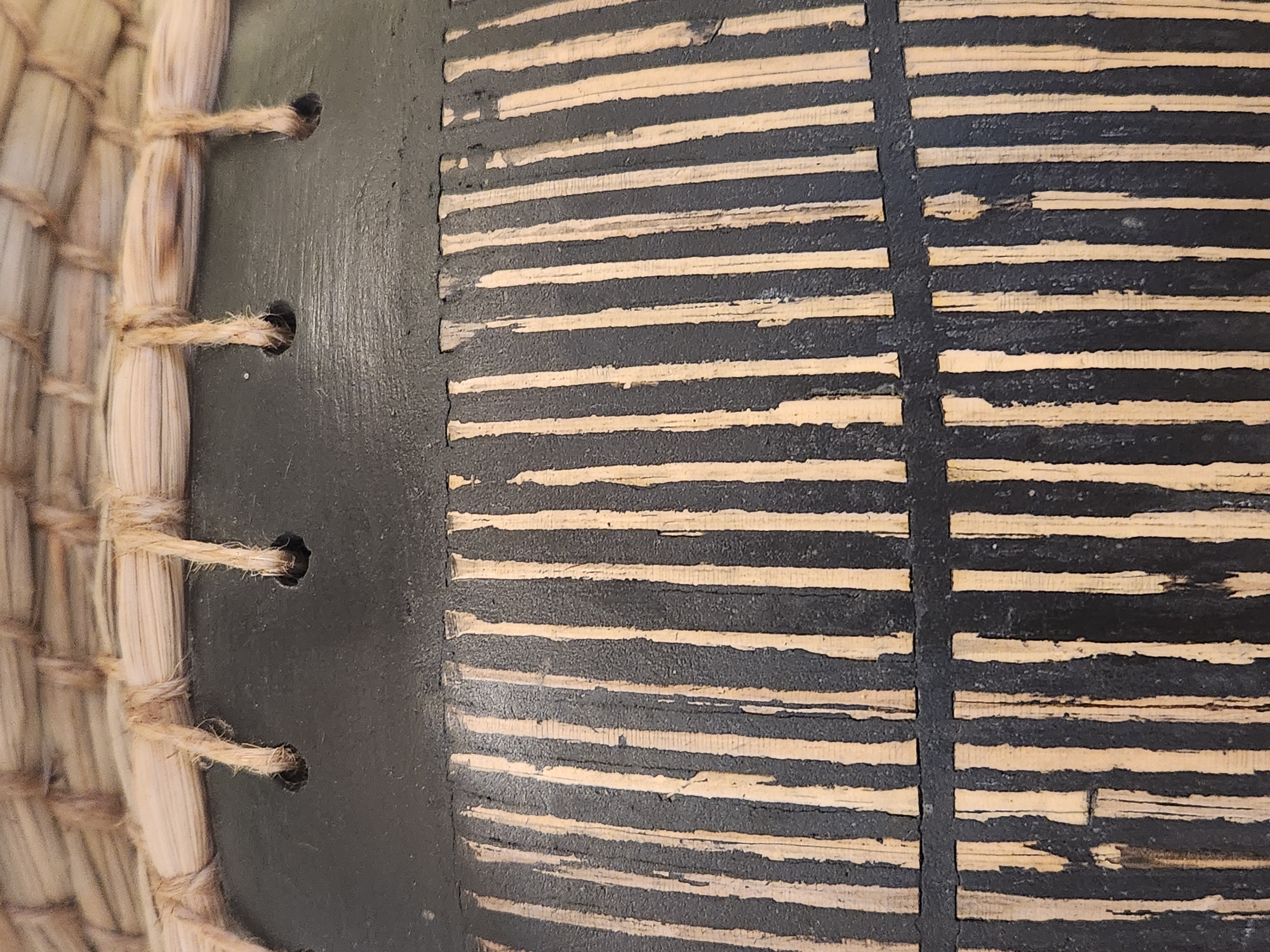 The image shows a close-up, top-down view of a textured surface, likely part of a basket or a decorative object. The top section features a woven material of light brown straw or reeds, with thicker strands running horizontally and thinner strands woven through them. Several holes are visible in this woven section, with rough, fraying ropes or strings passing through them and hanging down. Below the woven material, a band of matte black paint is applied. This black band is textured and appears somewhat rough. Below the black band, the surface transitions to a pattern of alternating vertical stripes of natural, light-colored wood or reed and the same matte black paint. The stripes are uneven in width and have a rustic, handmade appearance, with some areas of the black paint appearing chipped or worn away, revealing the lighter material beneath. The overall impression is one of handcrafted, rustic decoration. There is no information in the image that would indicate a specific location in the city of Maitland, United States of America.
