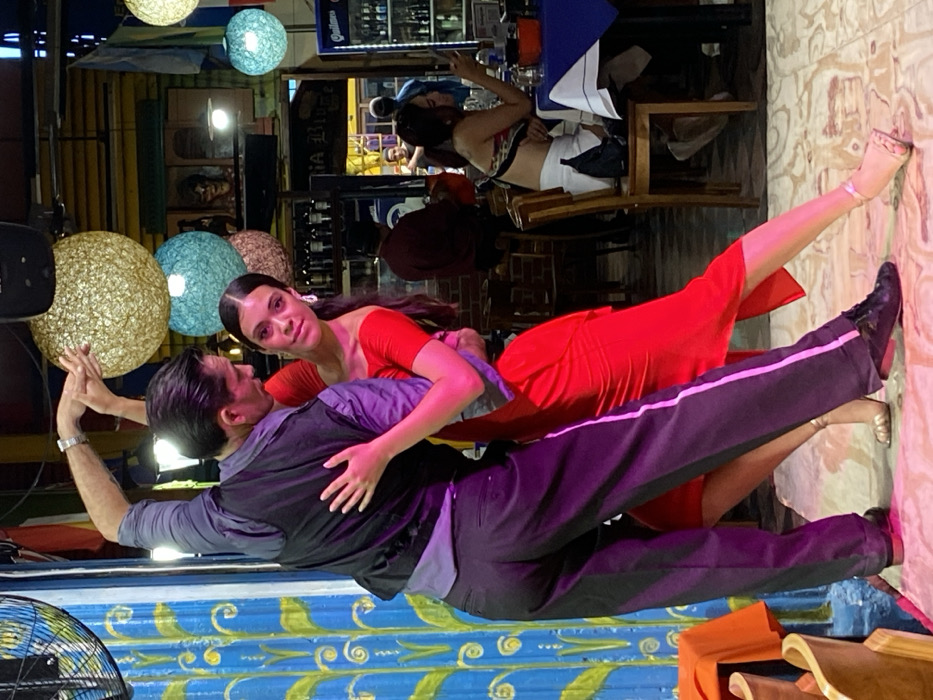 A man and woman are dancing in a brightly lit, colorful space. The woman is wearing a red dress and the man is in a dark suit with a stripe down the side. They are holding each other closely and looking at the camera. Round, decorative lanterns hang above them. Other people can be seen in the background, seated at tables. The floor is tiled with a patterned design.
