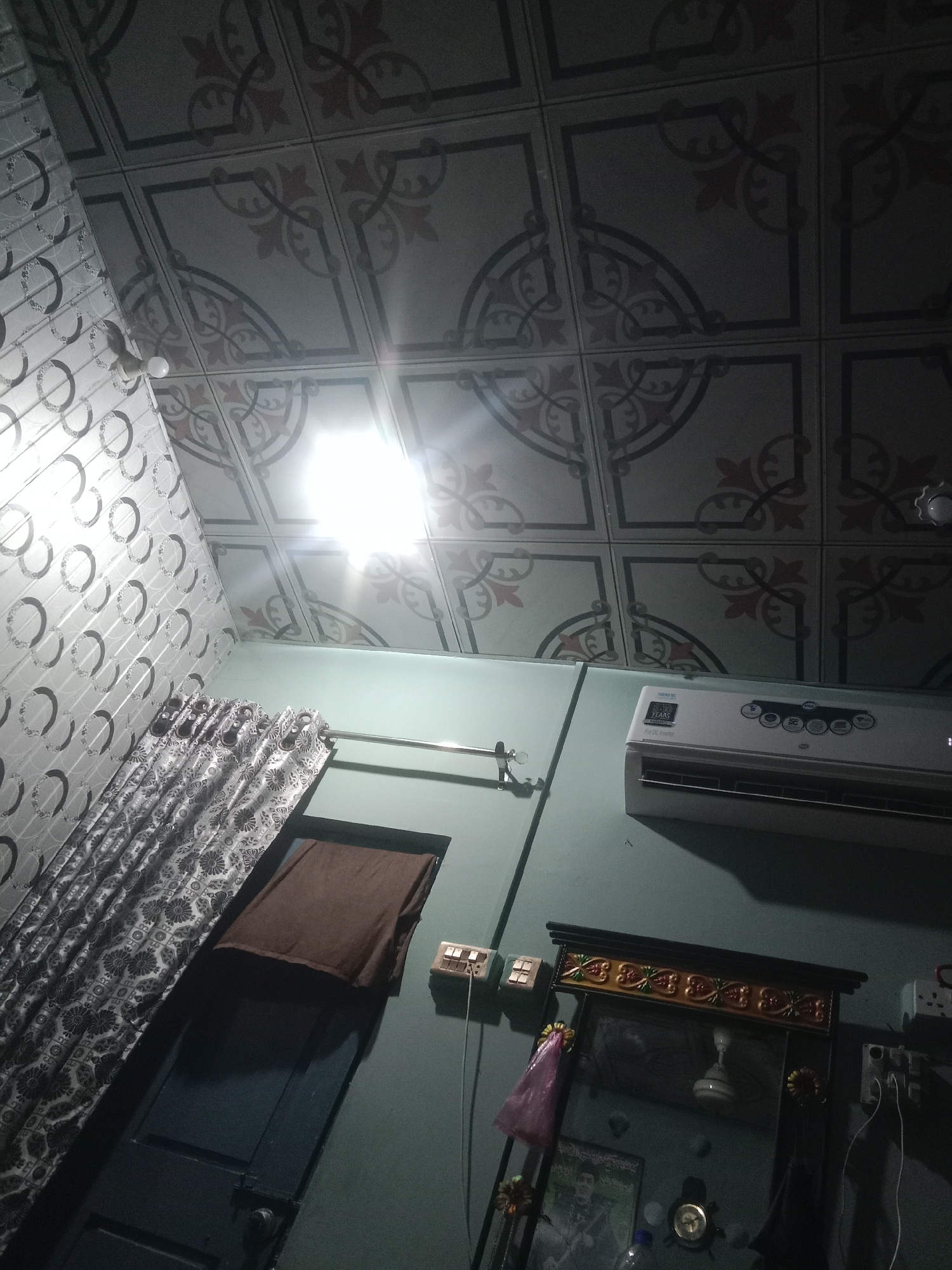 The image depicts an indoor scene, likely a room in Larkana, Pakistan. The ceiling is covered with patterned tiles. A bright overhead light illuminates the center of the frame, casting shadows. On the right, a white air conditioning unit is mounted on a light blue wall. Below it, a decorative mirror with an ornate frame hangs on the wall. Various objects are visible on the wall around the mirror, including electrical outlets and switches, a pink plastic bag, and what appears to be a framed picture. To the left, a window or doorway is partially covered by patterned curtains and a brown cloth. The overall impression is of a lived-in domestic space.