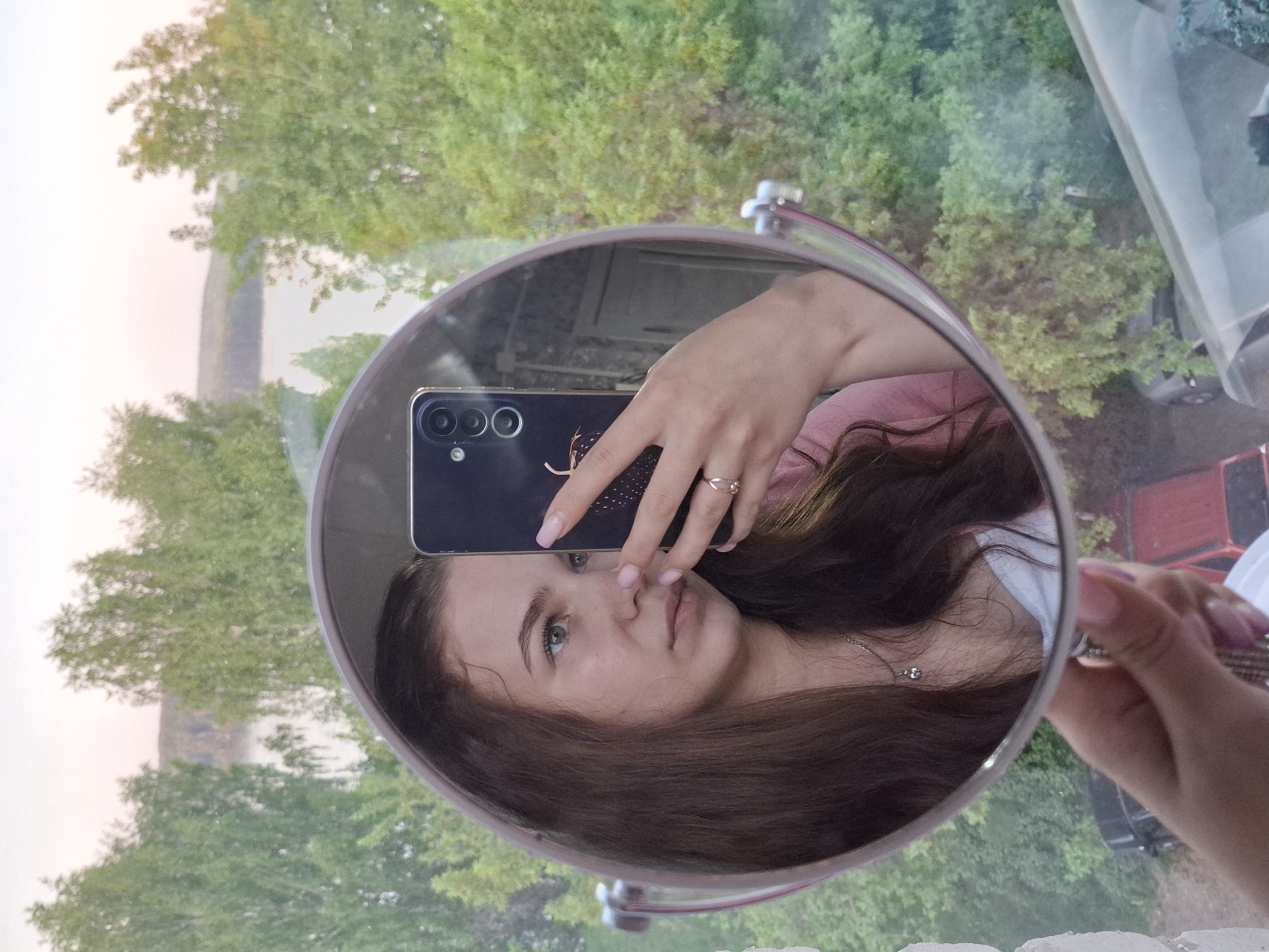 A young woman is taking a mirror selfie indoors, reflecting a view of the cityscape of Chaikovsky, Russia. The mirror is round and silver, held in her right hand. She is holding a black smartphone in her left hand, covering part of her face. She has fair skin, brown hair, and is wearing a ring. The reflection shows trees and buildings visible through the window, including a red car. The background suggests a residential area during daylight.