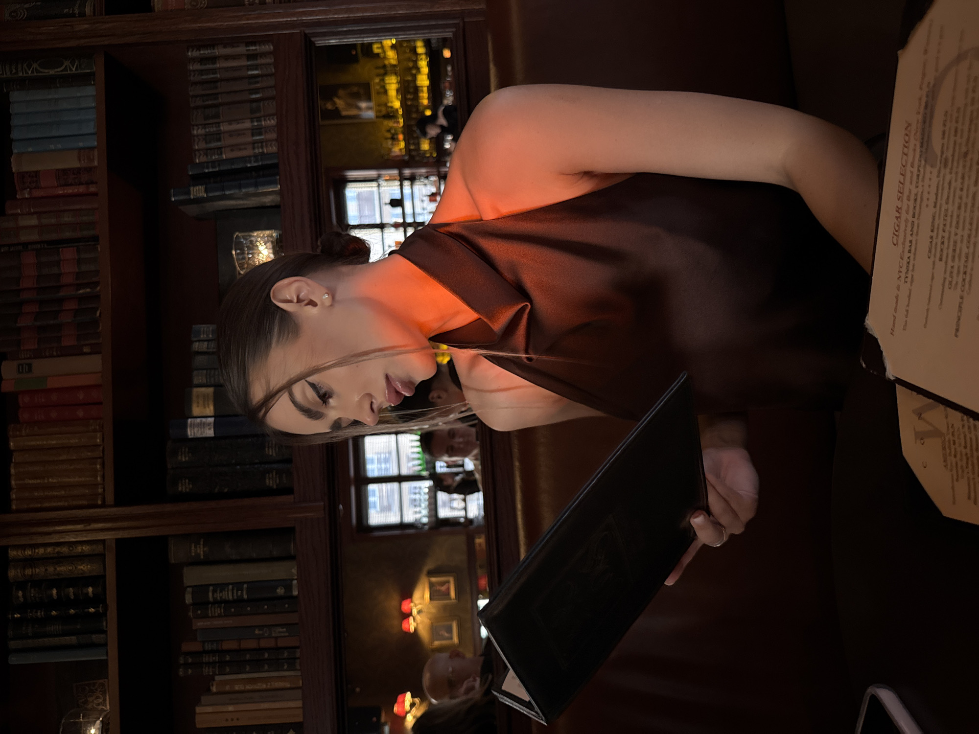 The image shows a woman in a dimly lit interior, possibly a restaurant or bar, evidenced by the presence of a cigar selection menu. The woman is wearing a dark top and is holding a menu. The background features a bookshelf filled with books. A reflection in a nearby surface shows glimpses of the interior, including lighting and other patrons. The location of the image is possibly the Tynska Bar and Books.