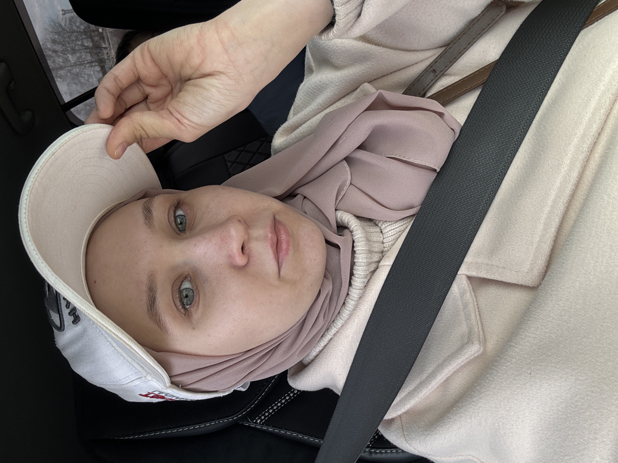 A young woman is sitting in the driver's seat of a car, wearing a white baseball cap, a hijab, and a coat. She is wearing a seatbelt. She is looking at the camera and holding the cap. The location is Chaikovsky, Russia.