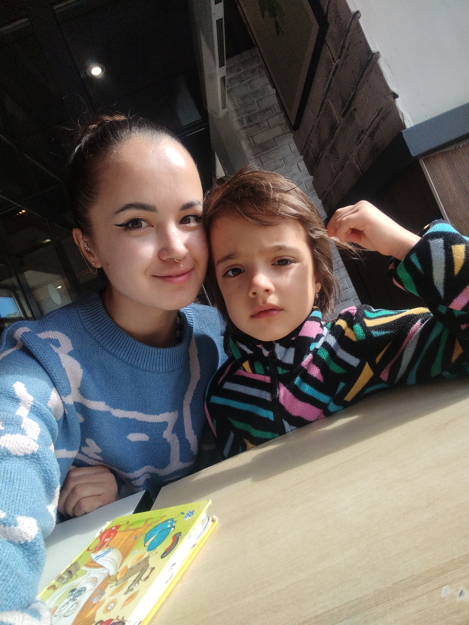 The image shows a woman and a child seated at a table. The woman has her dark hair pulled back in a bun, with visible multiple ear piercings and a nose ring. She is wearing a blue sweater with a white pattern. The child is wearing a multicolored patterned fleece jacket. The child is reaching up to touch their hair. A children's book is on the table in front of them. Based on visual features consistent with architectural details and items found in Chaikovsky, Russia, it is likely this photo was taken in that city.