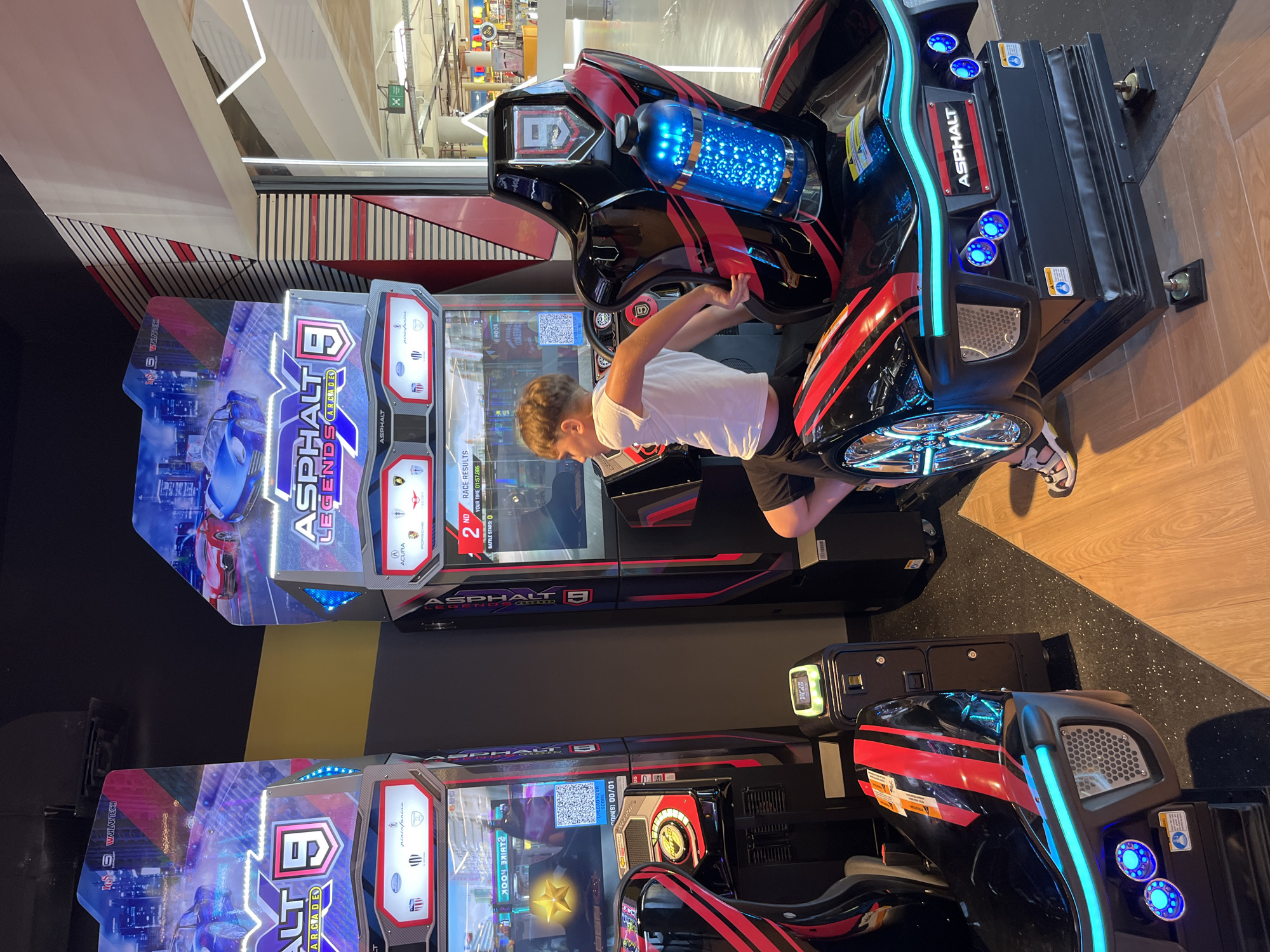 A young boy is playing an arcade racing game, "Asphalt 9: Legends," in a brightly lit entertainment center. He is seated in a realistic racing cabinet with a steering wheel and pedals, wearing a white t-shirt and dark shorts. The cabinet is designed to look like a sports car with black and red accents and vibrant blue LED lighting. The screen displays colorful graphics of cars and a racetrack.
To the boy's right, another "Asphalt 9: Legends" cabinet is visible, also featuring the game's branding and car imagery. The floor is a speckled black material transitioning to a wood-like finish. In the background, the ceiling features industrial lighting, and part of the entertainment center's infrastructure is visible, including metal beams and ductwork, suggesting an indoor public space. The overall atmosphere appears lively and engaging, typical of an arcade setting. The visible text on the game cabinet includes "ASPHALT 9 LEGENDS ARCADE," "WAVLOFTECH," and branding for car manufacturers like Acura, Porsche, and Lamborghini. The boy's posture suggests focus and enjoyment as he interacts with the game. The lighting indicates it is likely daytime, or the venue is well-lit regardless of the time.
