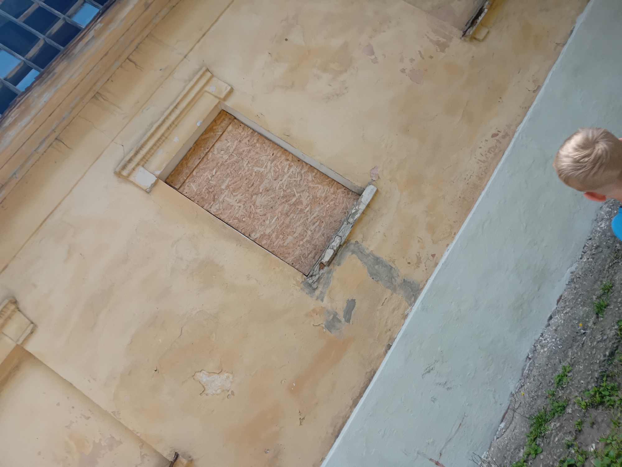 The image shows a close-up, angled view of a weathered building facade. The dominant color is a faded yellow, with patches of white plaster peeling away and darker staining indicating age and exposure. A boarded-up window, constructed from oriented strand board (OSB), is centrally positioned, set within a decorative beige frame. Above the boarded window, a portion of a multi-paned window is visible, with blue sky reflected in the glass. The lower part of the image features a light blue painted wall or section of the building, angled upwards. To the left of the blue wall, a gravelly area with sparse green vegetation is visible. A young child, with blonde hair, is partially visible in the bottom right corner, facing away from the camera. The overall impression is one of neglect or disrepair. The location is identified as Chuhuiv, Ukraine.