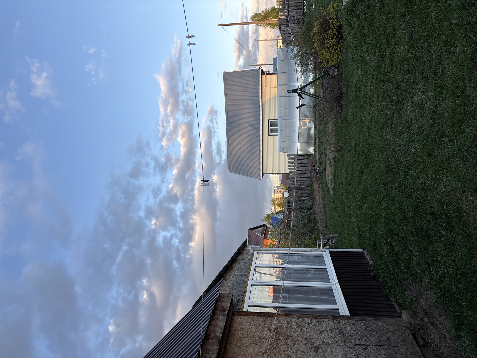 The image shows an exterior view of a residential area in Chaikovsky, Russia. In the foreground, there's a portion of a building with an unfinished exterior. A window is prominently featured with white frames, and the lower part of the building has brown siding. The roof is made of corrugated metal. Behind this building, there's another structure with light-colored siding and a gray metal roof. Next to this building is a greenhouse with a curved translucent roof. In the background, green grass and a wooden fence separate the properties. Overhead, there are utility lines with clothes pins attached. The sky is a mix of blue and gray clouds, suggesting either early morning or late evening. The lighting suggests it might be dusk or dawn.