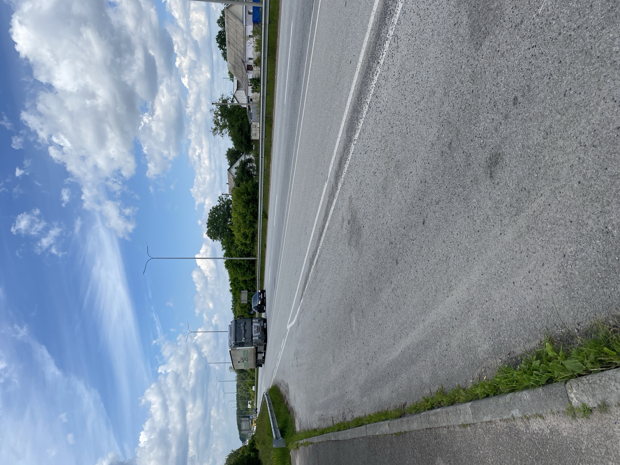 A two-lane road, with a concrete curb and grassy shoulder on the left, stretches into the distance under a partly cloudy blue sky. Several streetlights are visible along the roadside. On the right side of the road, a single dark-colored passenger car is traveling, followed by a dark-colored semi-truck with a white trailer. Beyond the road, a line of trees separates the highway from a residential area with several houses visible. The overall scene depicts a rural highway environment in Velyki Kosharyshcha, Ukraine, on a clear day.