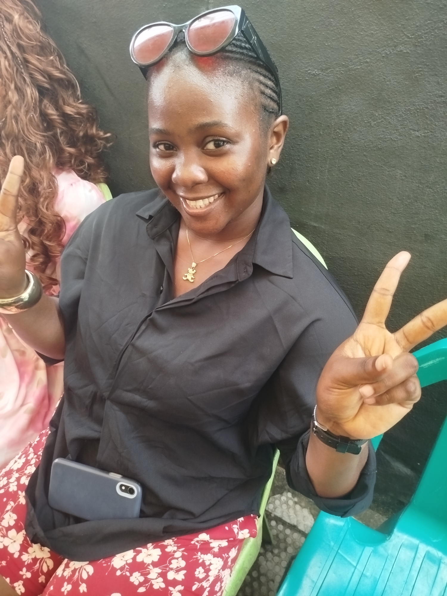 A Black woman with braided hair and sunglasses perched on her head smiles and makes a peace sign with her right hand. She is wearing a black button-up shirt, a gold necklace with a bear pendant, and a red skirt with a white floral pattern. A smartphone in a dark case is tucked into her shirt. She is sitting on a green chair outdoors, with a textured, dark green wall behind her. Another person with long, curly brown hair is partially visible to her left. The scene appears to be in Yenegoa, Nigeria.