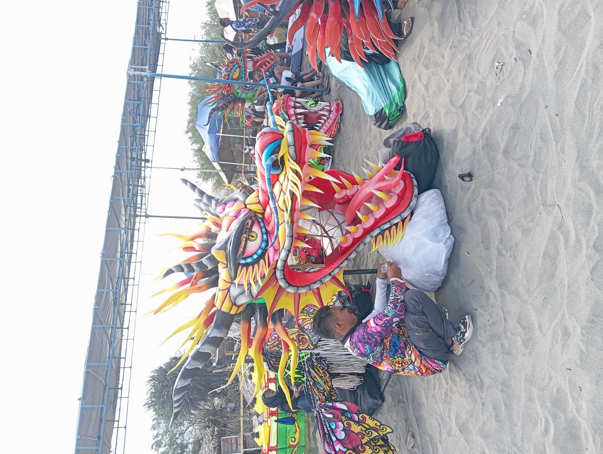 The image captures an outdoor scene on a sandy beach in Parangtritis, Indonesia, under a bright, overcast sky. The foreground is dominated by a large, vibrantly colored dragon head sculpture, painted in shades of red, yellow, blue, pink, and black, with sharp teeth and detailed eyes. A man in a colorful, patterned shirt and gray pants is crouching next to the dragon's head, appearing to be working on it, holding a small object in his hand. The dragon's body, adorned with intricate yellow and red flames, extends behind it. Several other dragon figures, some partially visible, are scattered around the scene. In the background, a blue tent is pitched, and people are gathered, some seated, suggesting a public event or preparation for one. A large, covered structure with a blue metal frame is visible in the upper portion of the image, possibly a temporary stage or shelter. The environment is characterized by sand dunes and sparse vegetation.