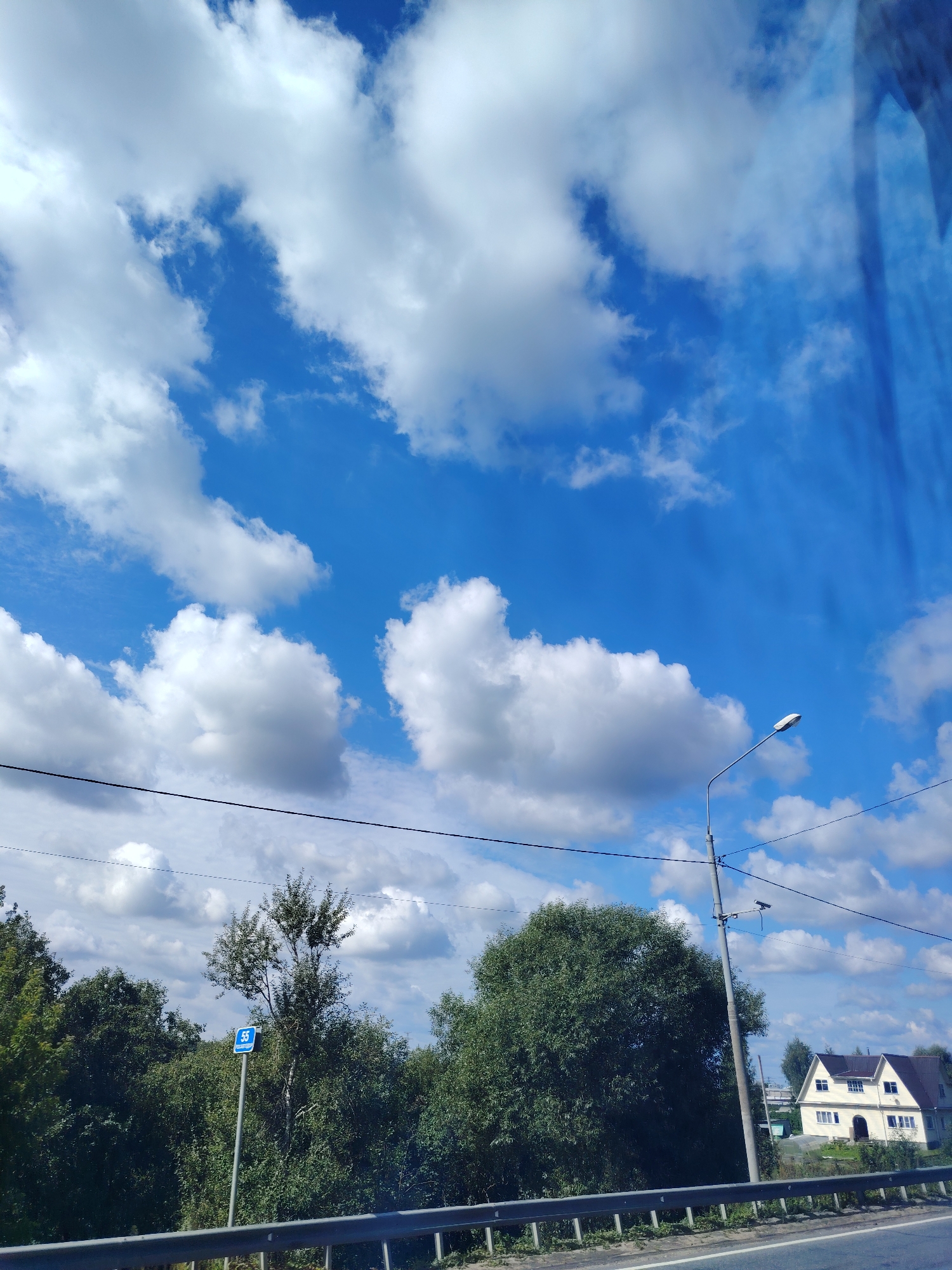 The image depicts a bright, sunny day with a clear blue sky dotted with fluffy white clouds. In the foreground, a road barrier runs horizontally across the bottom of the frame, suggesting a roadside view. Beyond the barrier, a dense line of green trees forms a natural backdrop. To the left, a blue sign with the number "55" is visible on a pole, likely a road marker. A tall street lamp stands to the right of the trees, its light fixture angled downwards. Further to the right, a two-story house with a dark roof and white walls is nestled among the greenery. The overall scene suggests a rural or suburban environment in the city of Peski, Russia.