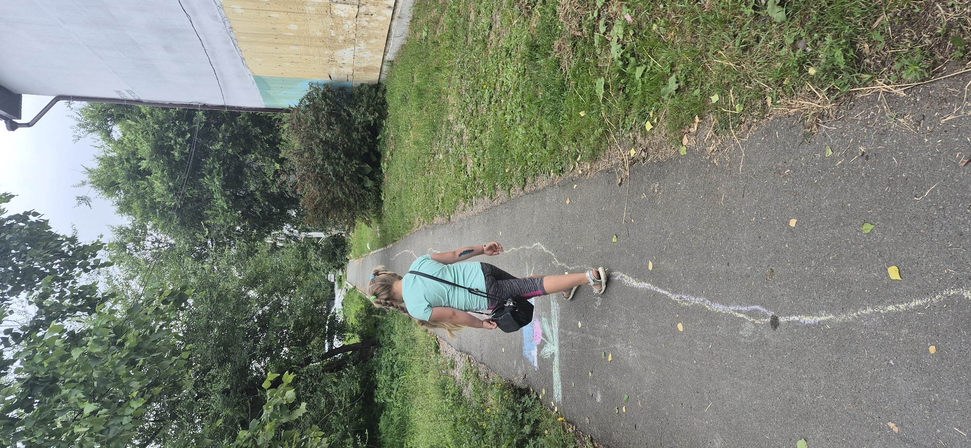 A full-shot, over-the-shoulder, outdoor vertical photo shows a young girl from behind walking on a paved path in an urban environment. The girl is blonde, with her hair in pigtails, and she is wearing a teal t-shirt and gray leggings with pink accents. She has a black purse slung over her shoulder. On the path, there are chalk drawings of flowers and a white chalk line running down the middle. The path is bordered by green grass and trees. A building with a light-colored wall is visible on the right side of the frame. The sky is overcast.