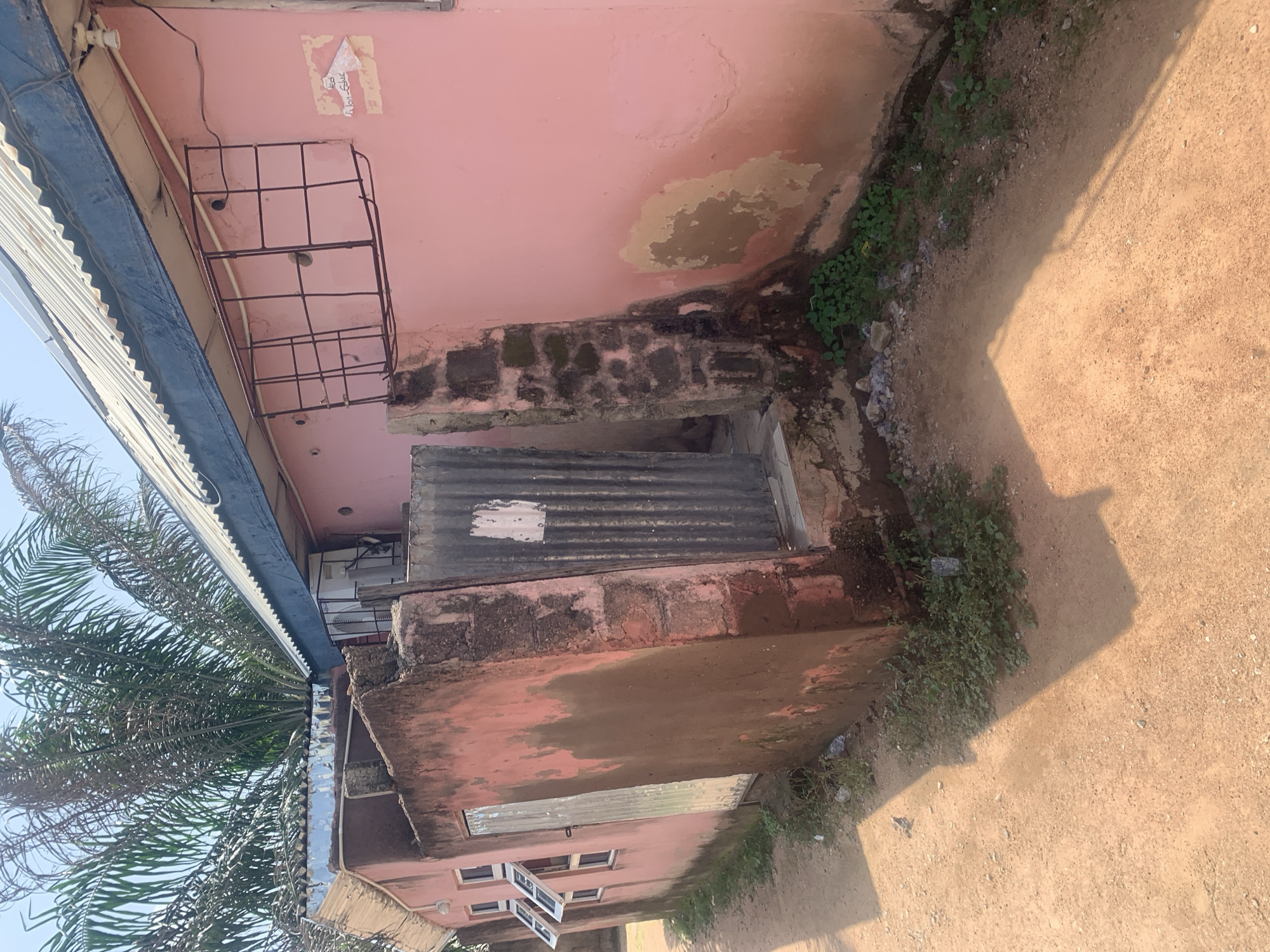 The image shows a pink, weathered building in Jalingo, Nigeria. The building has a corrugated metal roof and a blue fascia. The walls are stained and peeling in several places. There are several windows on the left side of the building, some of which are open. A metal door is visible on the left. A corrugated metal structure is attached to the right side of the building, adjacent to a brick column. An air conditioning unit is mounted on the wall. A metal grate covers a window or opening on the upper right side of the building. A piece of paper with text is affixed to the wall to the right of the grate. Palm fronds from a tree are visible at the top of the image. The ground in front of the building is a dirt path. The scene appears to be in a residential or commercial area.