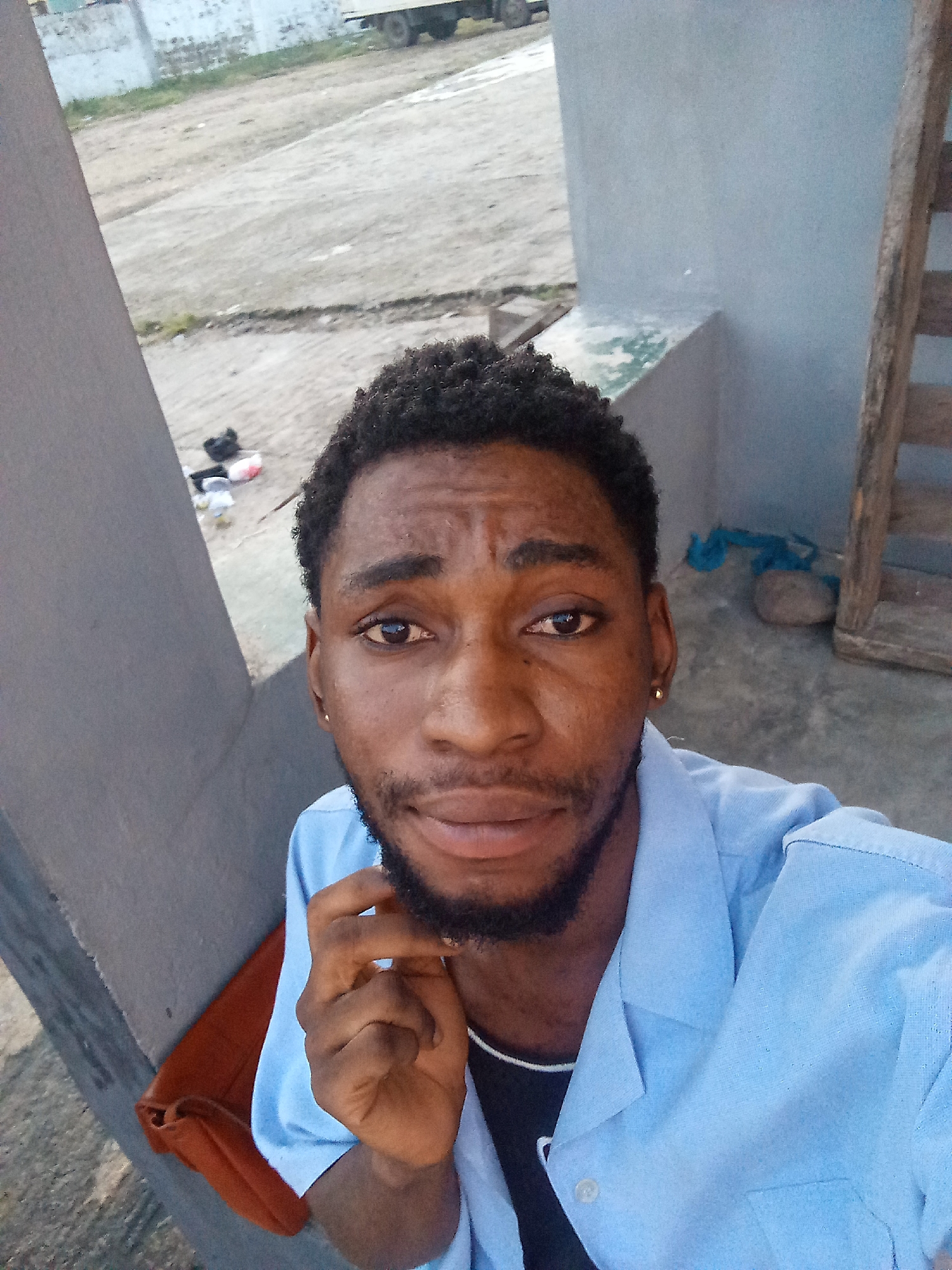 The image is a close-up, selfie-style photograph of a young Black man looking at the camera. He has dark, curly hair and a beard. He is wearing a light blue collared shirt over a dark shirt, with a silver chain visible. His right hand is raised to his chin. The background appears to be an outdoor, somewhat unkempt area. To the left, there is a light gray concrete pillar. Behind him, there is a concrete ground with some debris and a small pile of what appears to be trash. Further in the background, there is a light-colored structure, possibly a building or a wall, and a glimpse of a vehicle. To the right, a wooden structure with slats is visible, possibly part of a staircase or fence. The overall impression is a casual, everyday setting. The location is identified as the city of Lokoja, Nigeria.