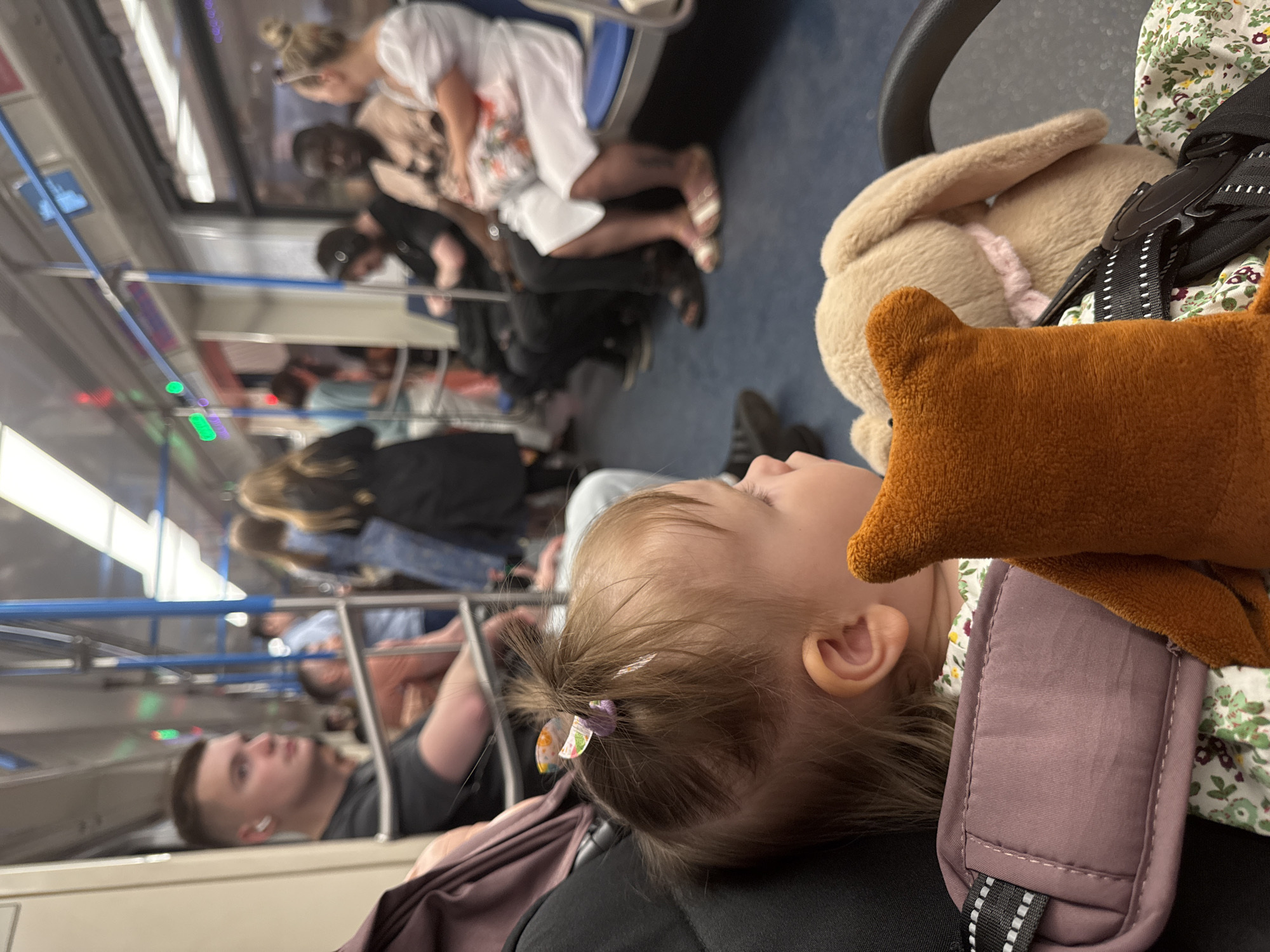 The image depicts the interior of a subway train, presumably in Moscow, Russia, given the visual cues of the subway car design and typical commuter attire. A young child, likely a toddler, is sleeping. A stuffed bunny and a brown bear plush are nearby. The child is seated in a carrier. Passengers are visible in the background, some seated and some standing. The overall atmosphere suggests a crowded subway during what appears to be a commute or travel period.