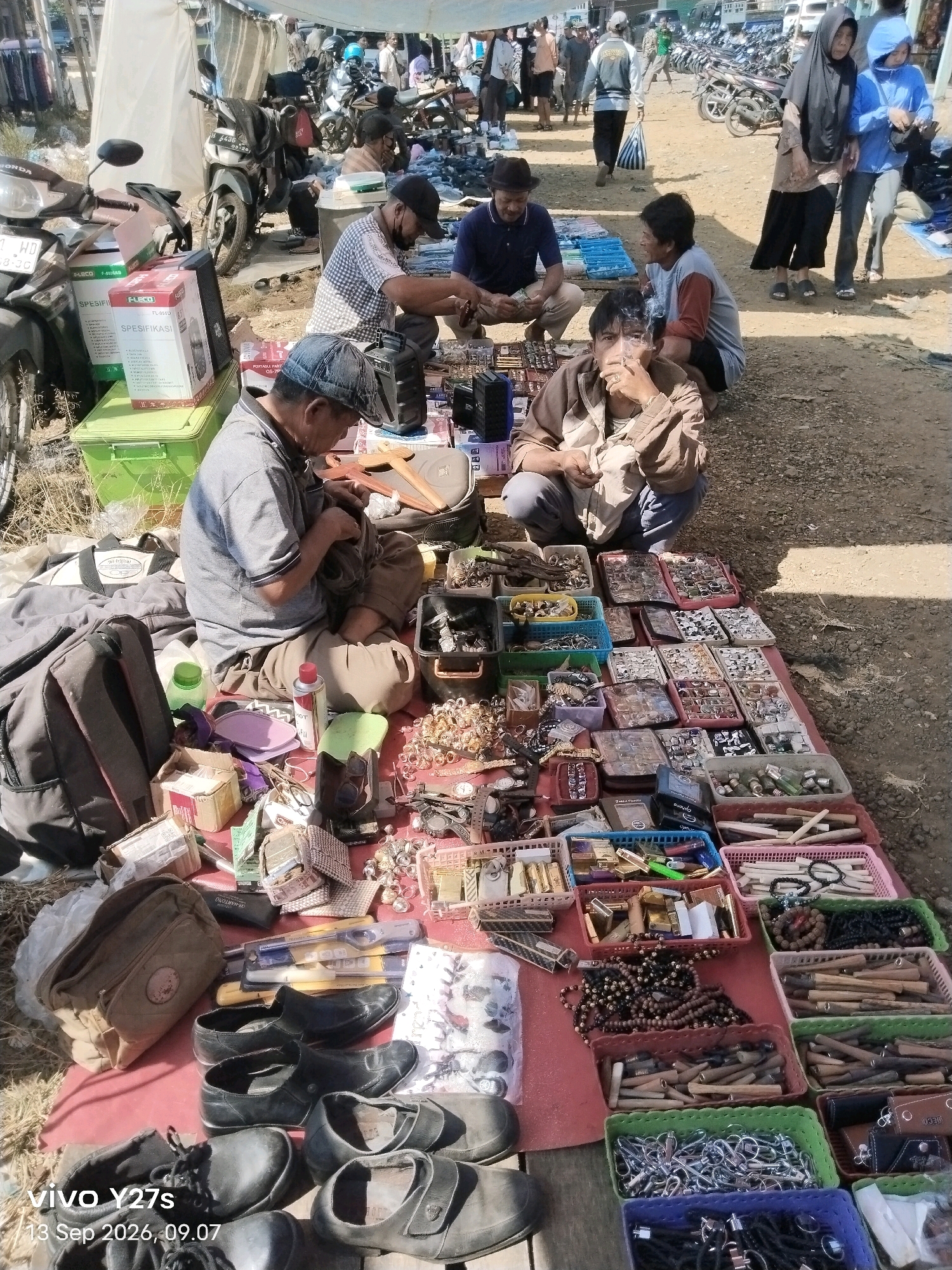 The image depicts a bustling outdoor market scene, likely in the city of Wonosido, Indonesia. The ground is covered with a dirt and gravel surface, and several people are seen engaged in commerce. In the foreground, a man wearing a black cap and a grey t-shirt is seated on the ground, appearing to be sorting through items. Beside him, a pile of black shoes and a brown bag are visible. Various stalls display an assortment of goods, including what appear to be jewelry, accessories, and possibly electronic items. In the background, motorcycles are parked, and other individuals are browsing or selling merchandise. The overall atmosphere suggests a typical day at a local market, with a focus on transactional activities.
