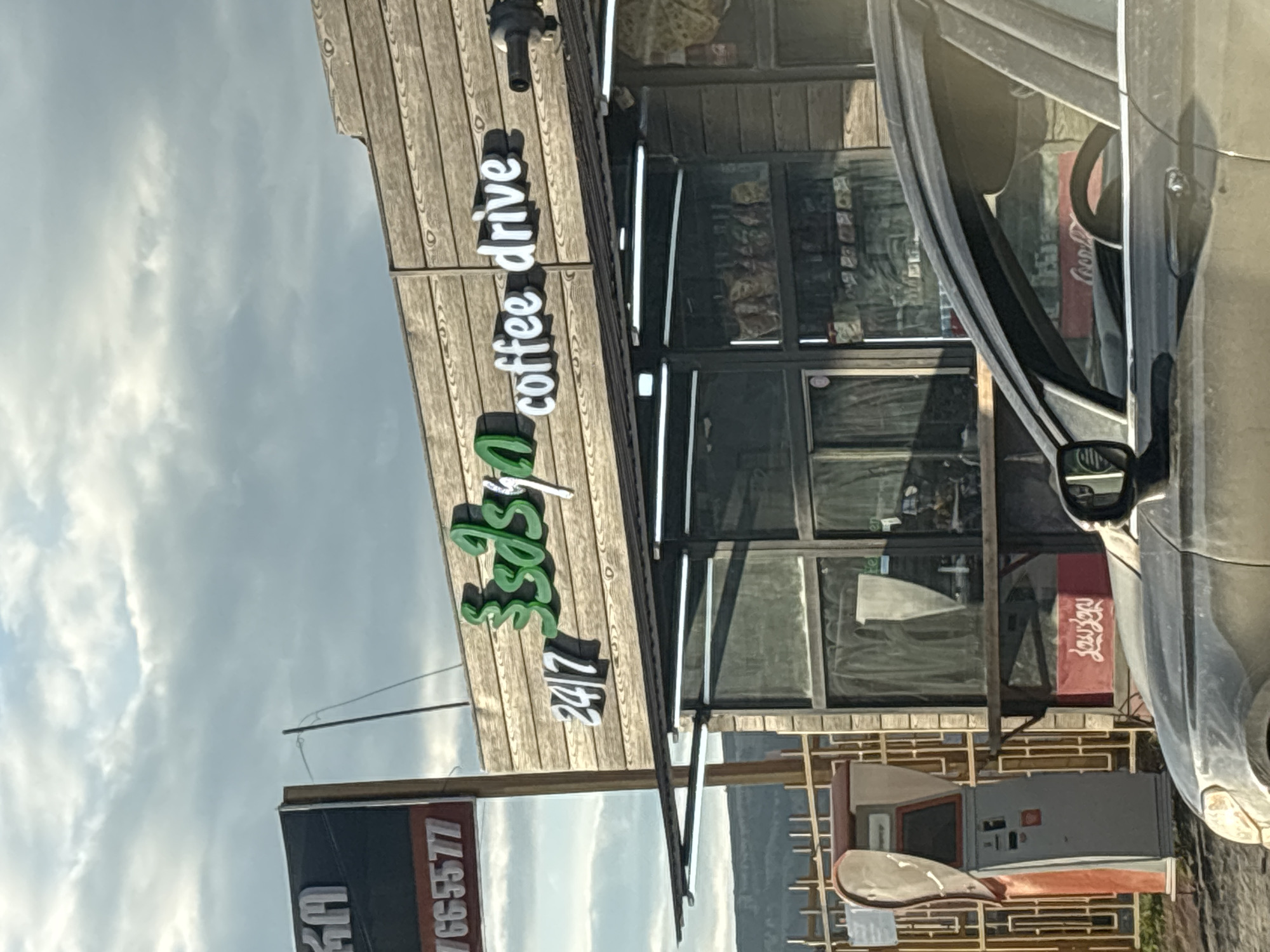 The image shows a "3S 24/7 coffee drive" business in Tbilisi, Georgia. The sign is mounted on a wooden structure. The storefront has glass windows, and a car is parked nearby. There's a gas pump visible. A phone number is displayed on a sign. The sky is cloudy.