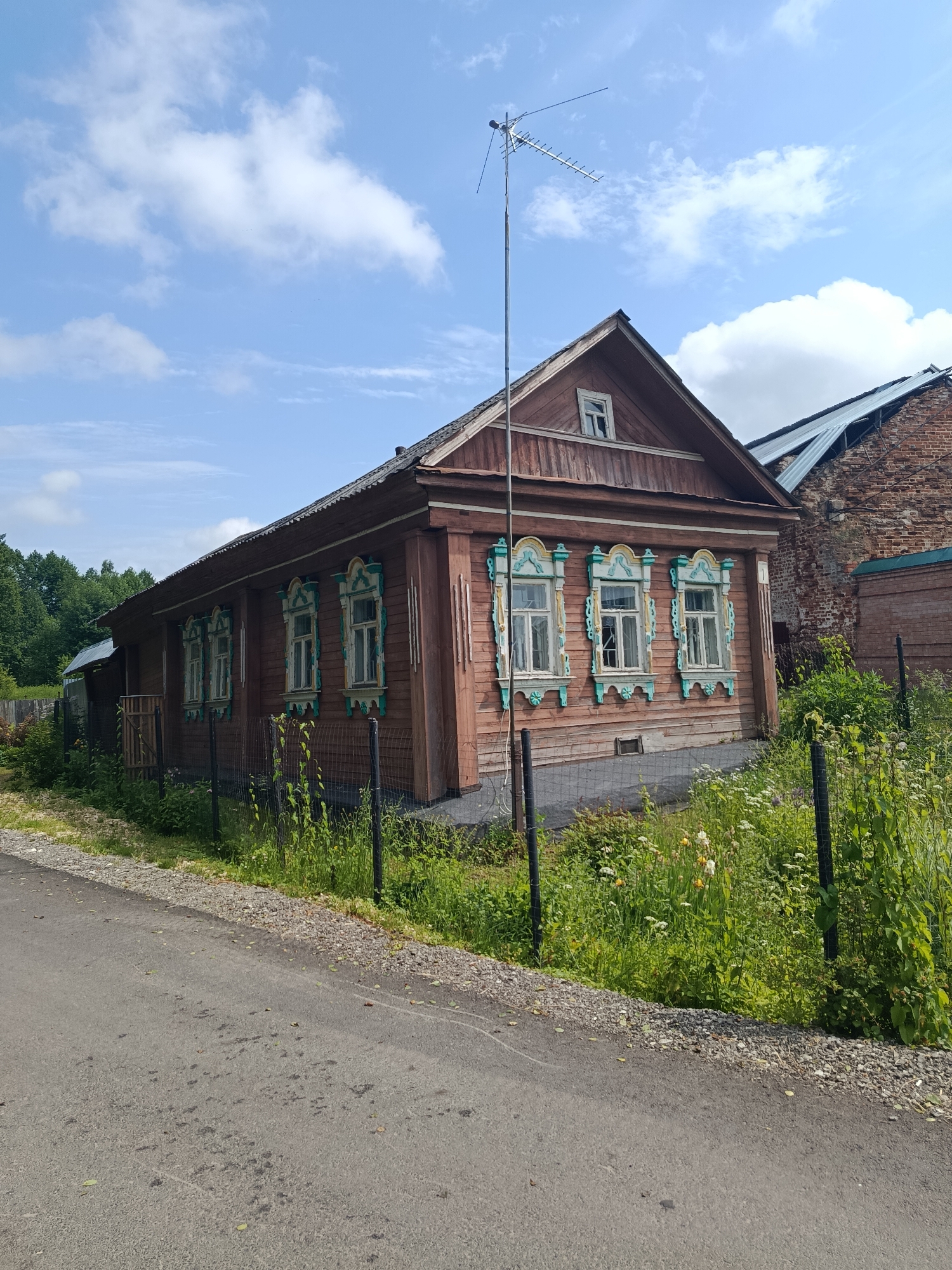 This is a medium-shot, eye-level, outdoor, daytime photograph taken in Radovitsy, Russia. The image features a traditional wooden Russian house with ornate window frames painted in turquoise and yellow. The house is situated on a slight incline, with a dark asphalt road in the foreground. A black wire fence runs along the property line, separating the house from the road and a patch of wild grass and wildflowers. To the right of the wooden house, a partially visible brick building with a metal roof is present, suggesting construction or renovation. A tall antenna stands in front of the wooden house, its reflection visible in one of the windows. The sky is blue with scattered white clouds. The environment appears rural or suburban, with trees visible in the background.