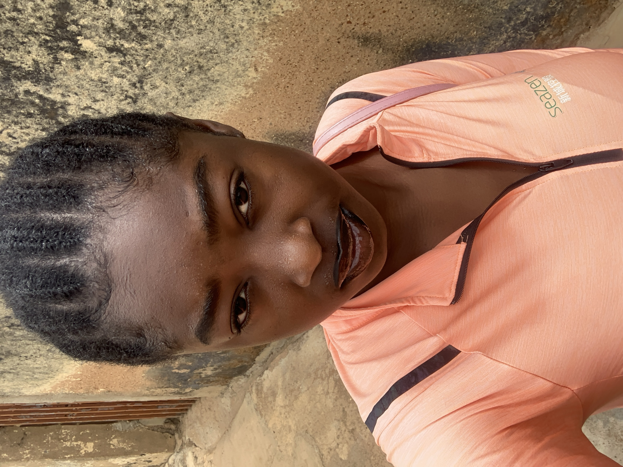 A young Black woman is shown in a close-up, eye-level shot. Her hair is braided and she is wearing a peach-colored zip-up jacket with black stripes. The location appears to be Rafin Par, Nigeria, as indicated by the prompt. The background consists of a weathered concrete surface. The woman's expression is neutral as she looks at the camera.