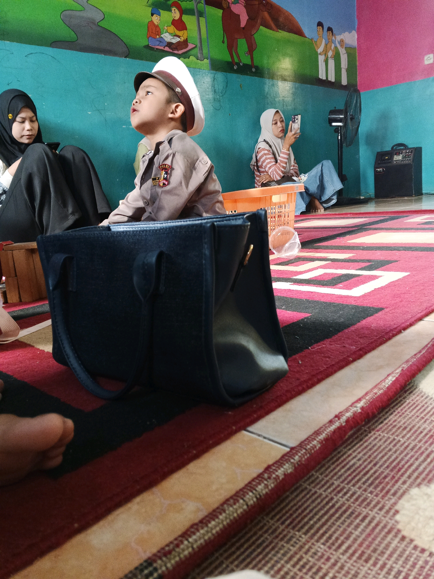 In an indoor setting, possibly a community space or home in Warungkiara, Indonesia, three individuals are present. A child, appearing to be around 5-7 years old, is seated and dressed in a brown uniform with a white cap, looking upwards. To the child's left, a woman wearing a black hijab and black clothing is seated, looking down at a smartphone. To the right of the child, another woman wearing a light-colored hijab and a striped shirt is seated, holding a smartphone. In the foreground, a dark blue handbag is placed on a red and white patterned rug. The background features a wall with colorful murals depicting people and animals, and a black standing fan is visible. The scene appears to be informal, with individuals engaged with their devices or observing something.