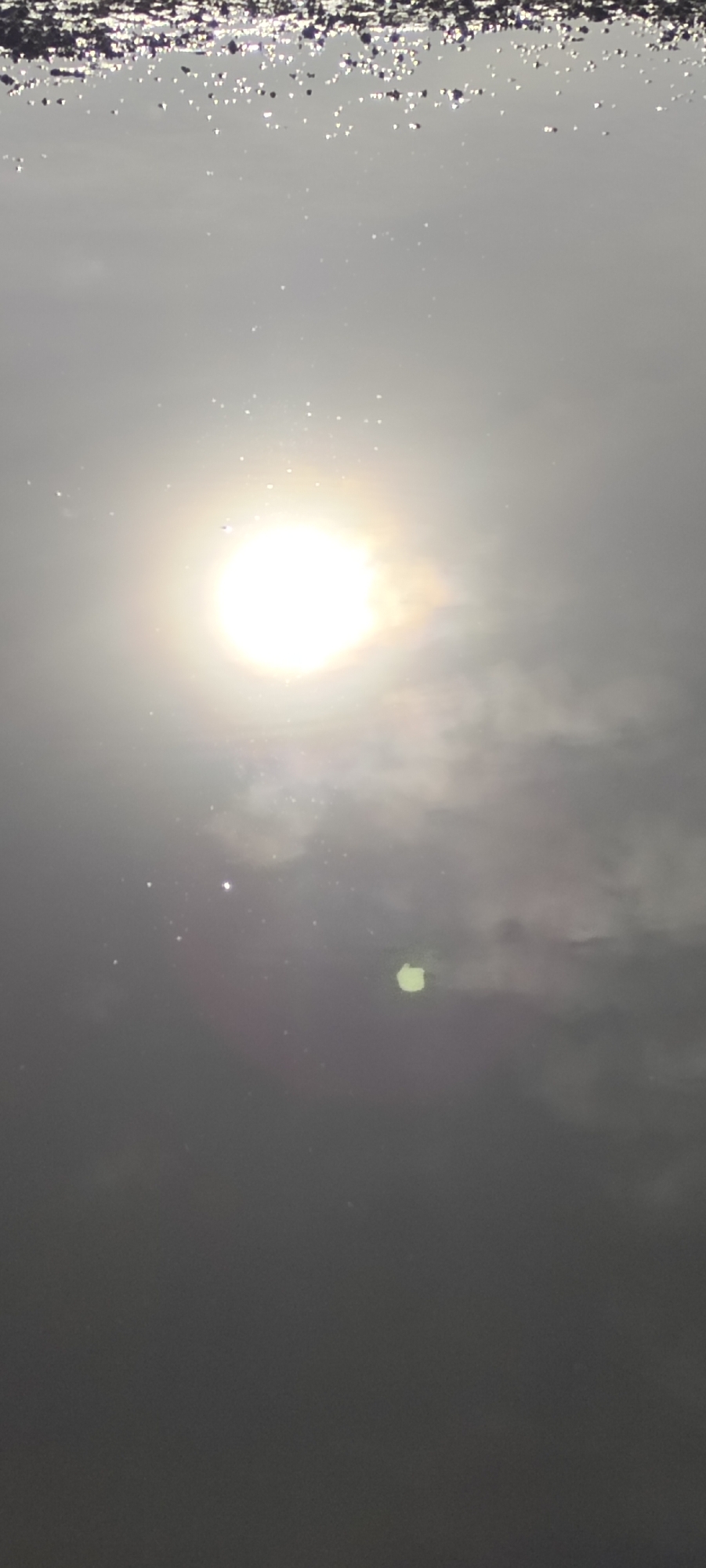 The image depicts a bright sun obscured by a thin layer of clouds, as seen from Lhokseumawe, Indonesia. The top portion of the image shows a dark, jagged edge, suggestive of the shoreline or debris floating on the water's surface. The sun is the focal point, shining brightly through the hazy atmosphere, and its light is partially diffused by the surrounding clouds. The lower part of the image fades into a dark, undifferentiated area, potentially representing the ocean's depths or a shaded foreground. There are no visible people, subjects, objects, actions, or interactions, beyond the depiction of the sun, clouds, and water.