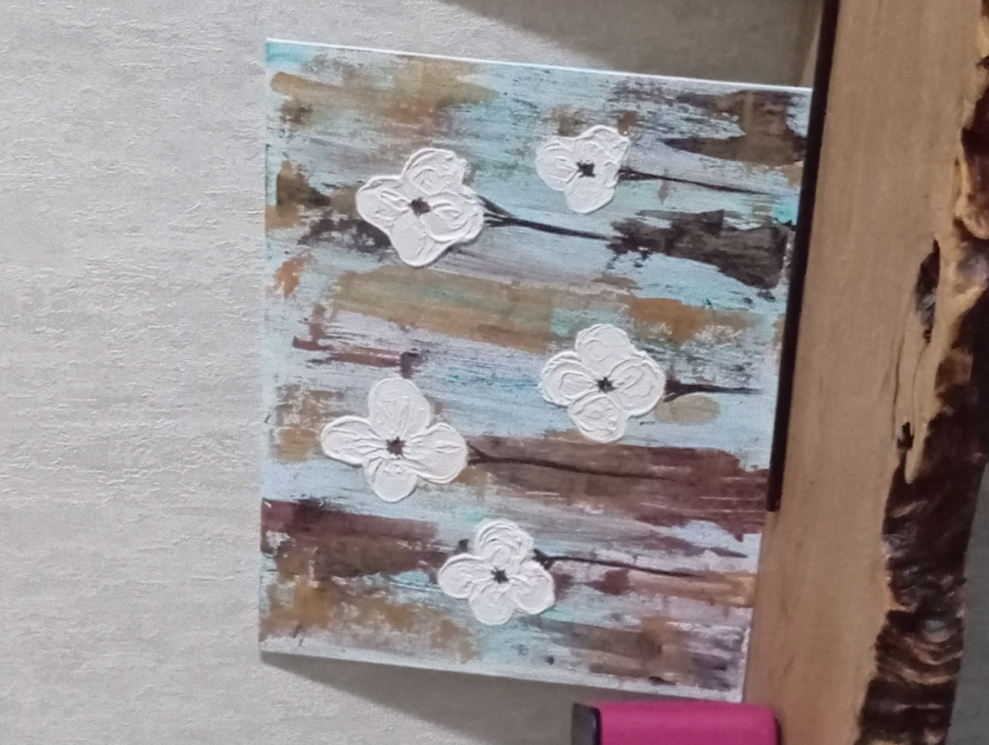 The image shows a textured painting of five white flowers with dark centers, rendered in a impasto style, displayed on a wooden surface against a textured wall. The painting features a background of abstract brushstrokes in shades of brown, white, and light blue. A bright pink object is partially visible on the left side of the frame. The scene is likely indoors, with the painting positioned as decorative art. The image does not contain any information that can be used to determine its location in Yekaterinburg, Russia.