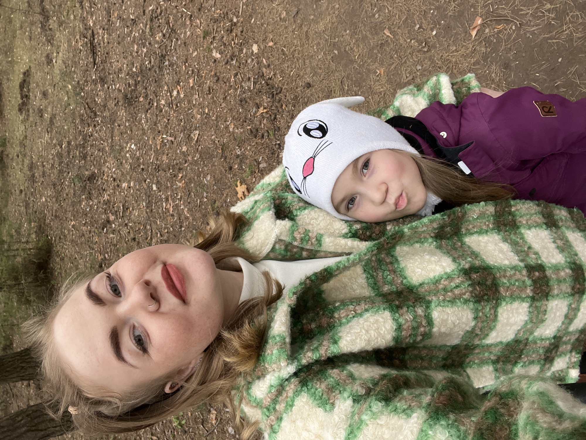 This media file captures an adult female and a young child lying on the ground outdoors in Chaikovsky, Russia. The image is taken from an elevated perspective, looking down on the subjects, who appear to be relaxed on their backs.
The setting is a natural environment, likely a park or wooded area, with the ground covered in brown dirt, dry leaves, and some sparse green patches. The overall atmosphere suggests a cool day, as both individuals are dressed in warm attire.
The adult female, with light blonde hair, is wearing a white long-sleeved top and a warm, shaggy plaid coat or blanket in shades of cream, green, and brown. She has subtle makeup, including a nose ring, and looks calmly towards the camera.
The young child is snuggled close, wearing a white knit hat designed as a bunny face with large black eyes and a pink nose. She has a dark purple winter jacket, on the sleeve of which is a brown patch featuring a stylized logo, possibly an "R" or similar symbol. The child's expression is a slight pout, as she also looks towards the camera. Both subjects appear at ease, possibly enjoying a quiet moment outdoors.