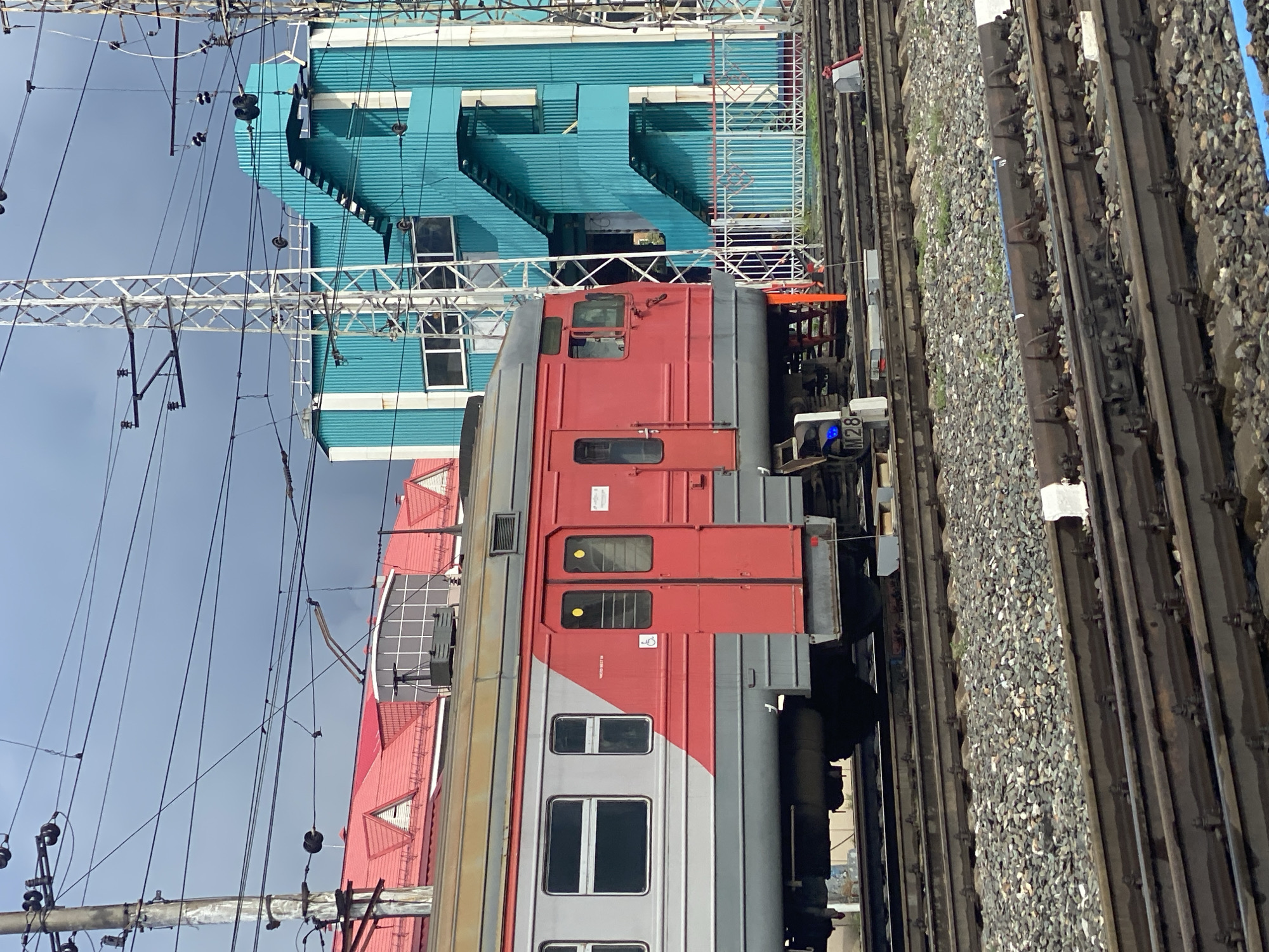 The image captures a view of a train at a station in Novokuznetsk, Russia, under an overcast sky. A red and gray passenger train is positioned on the tracks in the foreground. To the right of the train, a bright turquoise building with white accents and an external staircase is visible. Behind the train, a building with a red roof is partially obscured. The area is characterized by multiple sets of train tracks, ballast, and overhead electrical wires with supporting poles and towers. A blue signal light is illuminated on a marker post near the train. There are no people visible in the image. The environment suggests an active railway operational area.