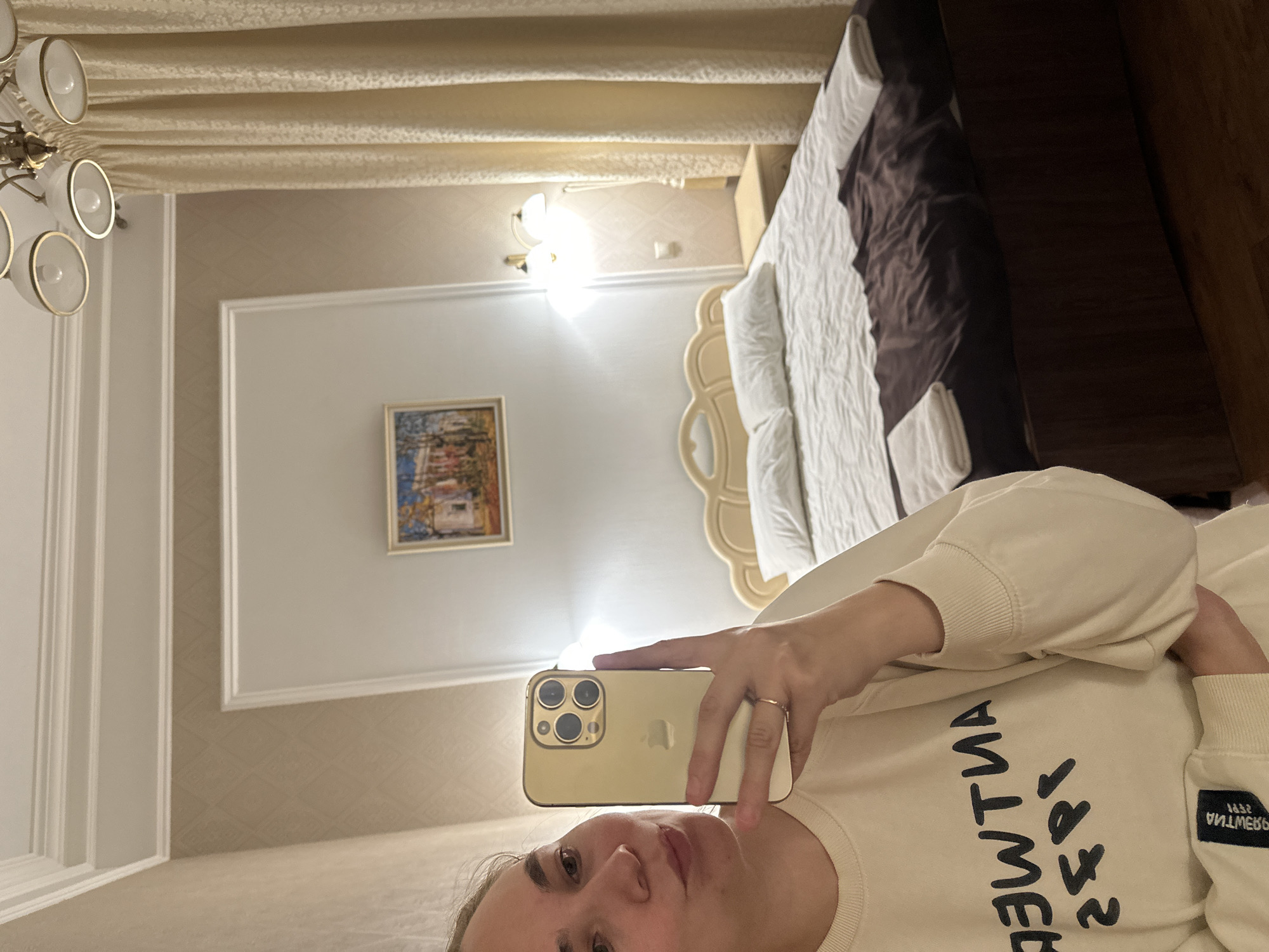 A young woman with light-colored hair is taking a selfie in a hotel room. She is wearing a light-colored sweatshirt with black text on it. She is holding a gold iPhone in her right hand. In the background, there is a bed with white linens and a dark brown comforter. There are two white towels folded on the bed. Above the bed, there is a headboard. On the wall, there is a framed picture and a wall lamp. To the right of the bed, there is a bedside table and a cream-colored curtain. The room has beige wallpaper with a diamond pattern. A chandelier is visible on the ceiling. The image appears to have been taken in Khvalynsk, Russia, likely in a hotel room.