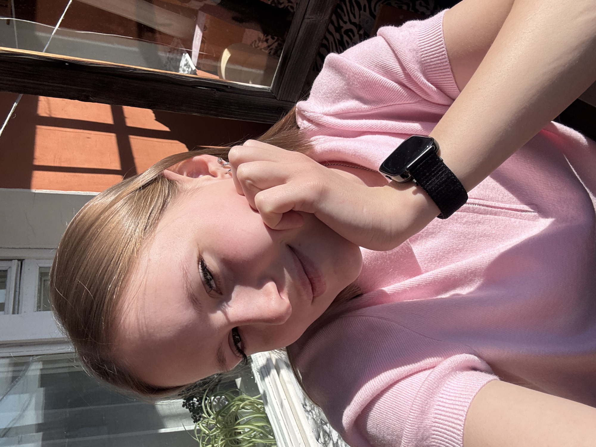 A close-up, tilted photograph shows a young woman with light brown hair wearing a pink sweater and a black smartwatch on her wrist. She is touching her face with her hand. Behind her are windows, an exterior wall painted reddish-brown, and a window frame. Outside the window, greenery is visible. The photograph appears to have been taken outdoors in daylight. There is no definitive information in the image to identify that the location is Chaikovsky, Russia.