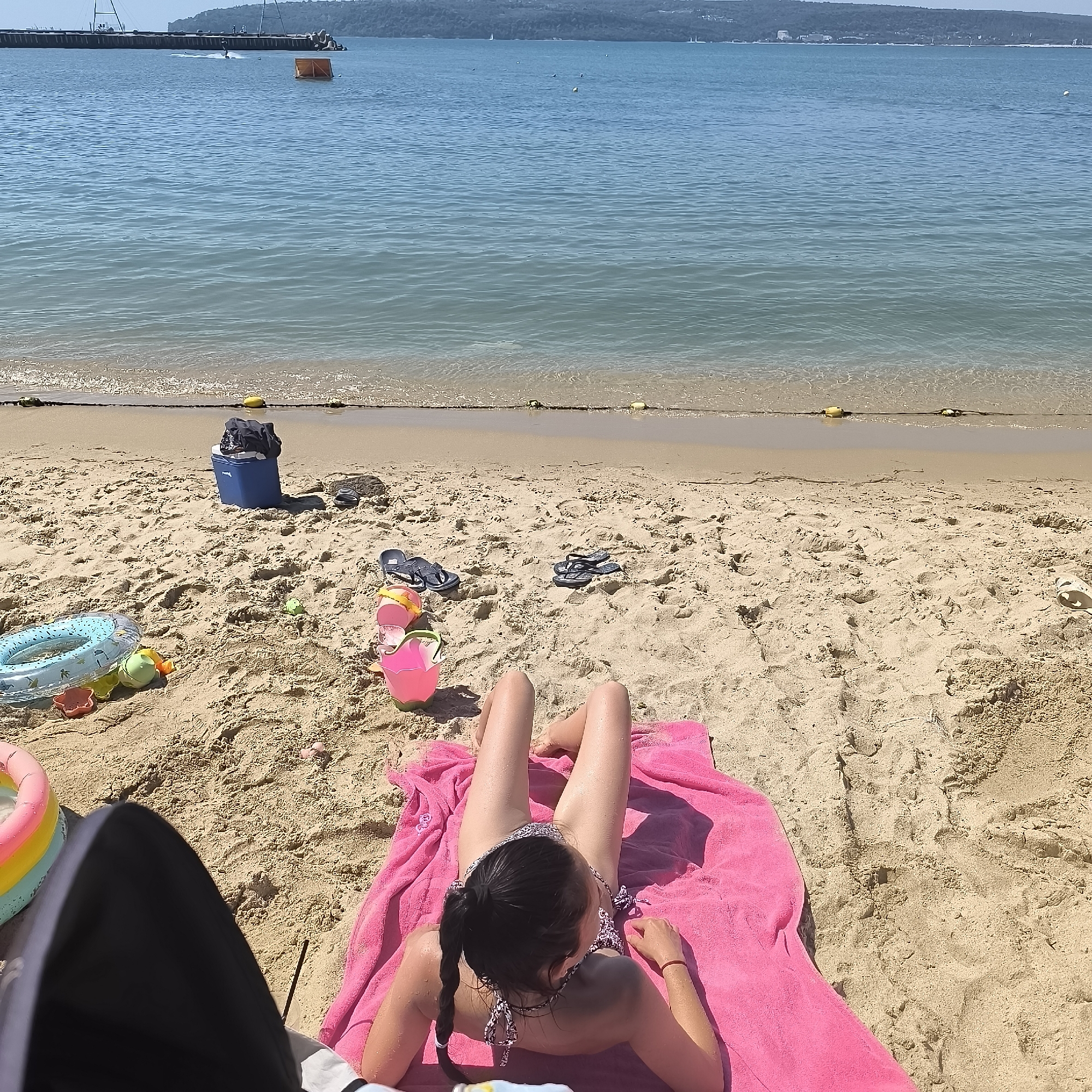 This image depicts a beach scene in Varna, Bulgaria, under clear, sunny skies, suggesting it's midday. The primary subject is a young girl lying on a bright pink towel on the sand, facing away from the camera and towards the sea. She is wearing a bikini and has her hair in a braid. Her legs are stretched out in front of her. The ocean is calm and blue, stretching to the horizon where a hazy, green hillside with some buildings is visible. A long pier extends into the water from the left, with a person on a jet ski visible near it, and a floating orange marker buoy in the water.
Scattered on the sand around the girl are various beach toys, including a blue inflatable ring with a pattern, several colorful plastic sand molds, and a pink bucket. A blue cooler bag filled with dark items sits further up the beach, and a pair of black flip-flops are near it. Another pair of flip-flops is also visible closer to the center of the frame. The sand itself is golden and appears well-trodden, with some footprints visible. The overall atmosphere is one of relaxation and summer enjoyment. No visible text is present in the image.