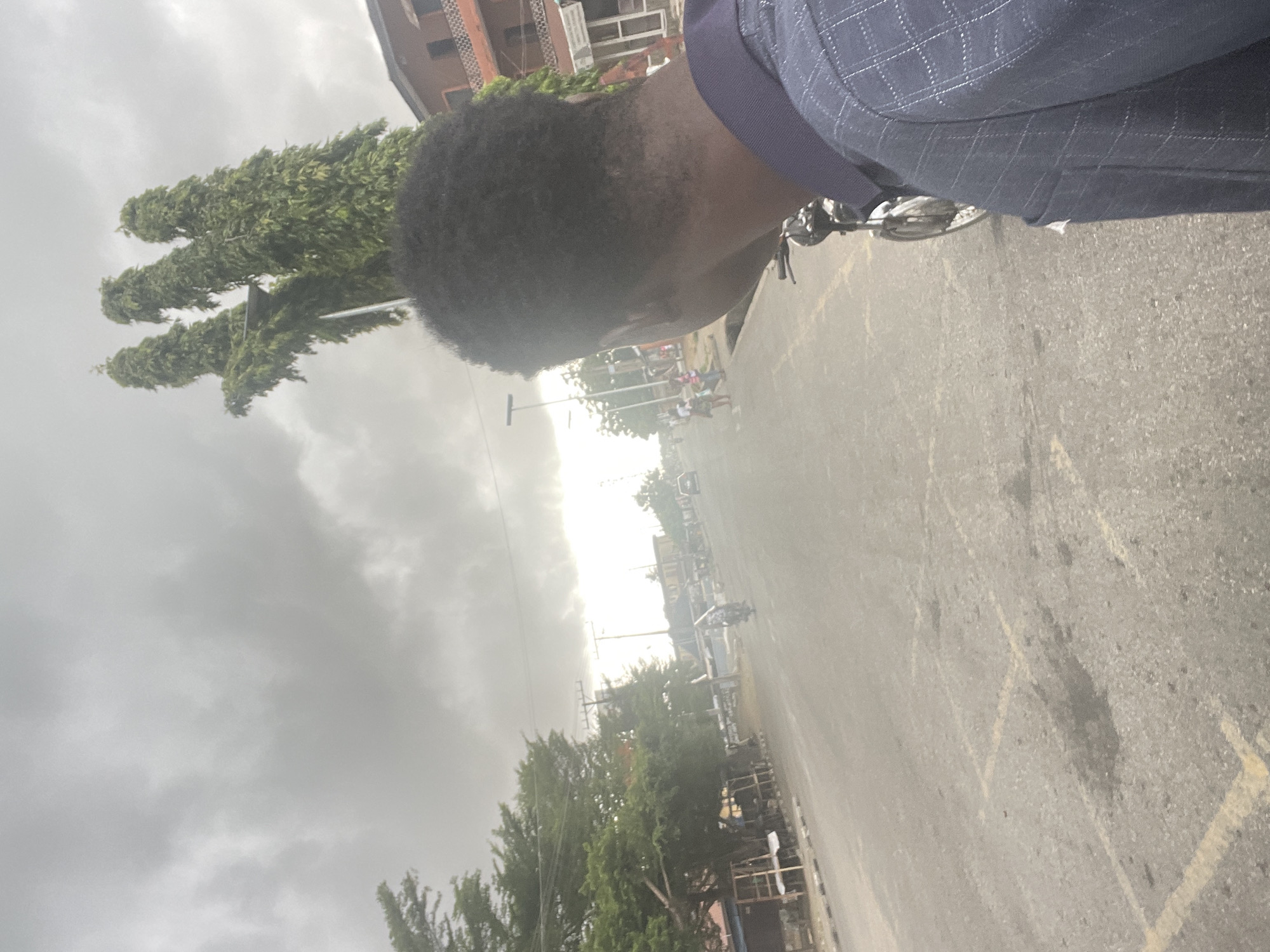 The image depicts a street scene in Badagry, Nigeria, under an overcast sky. The street runs horizontally, appearing light gray, with faded yellow parking lines on the surface. The side of a person’s head is visible in the foreground; they have dark hair and are wearing a dark blue checkered blazer. To the left of the frame are tall, thin trees with dense foliage. In the distance, along the street, are indistinct figures of people and vehicles; notably, a motorcycle. Buildings are faintly visible on either side of the street. The horizon is obscured by haze.