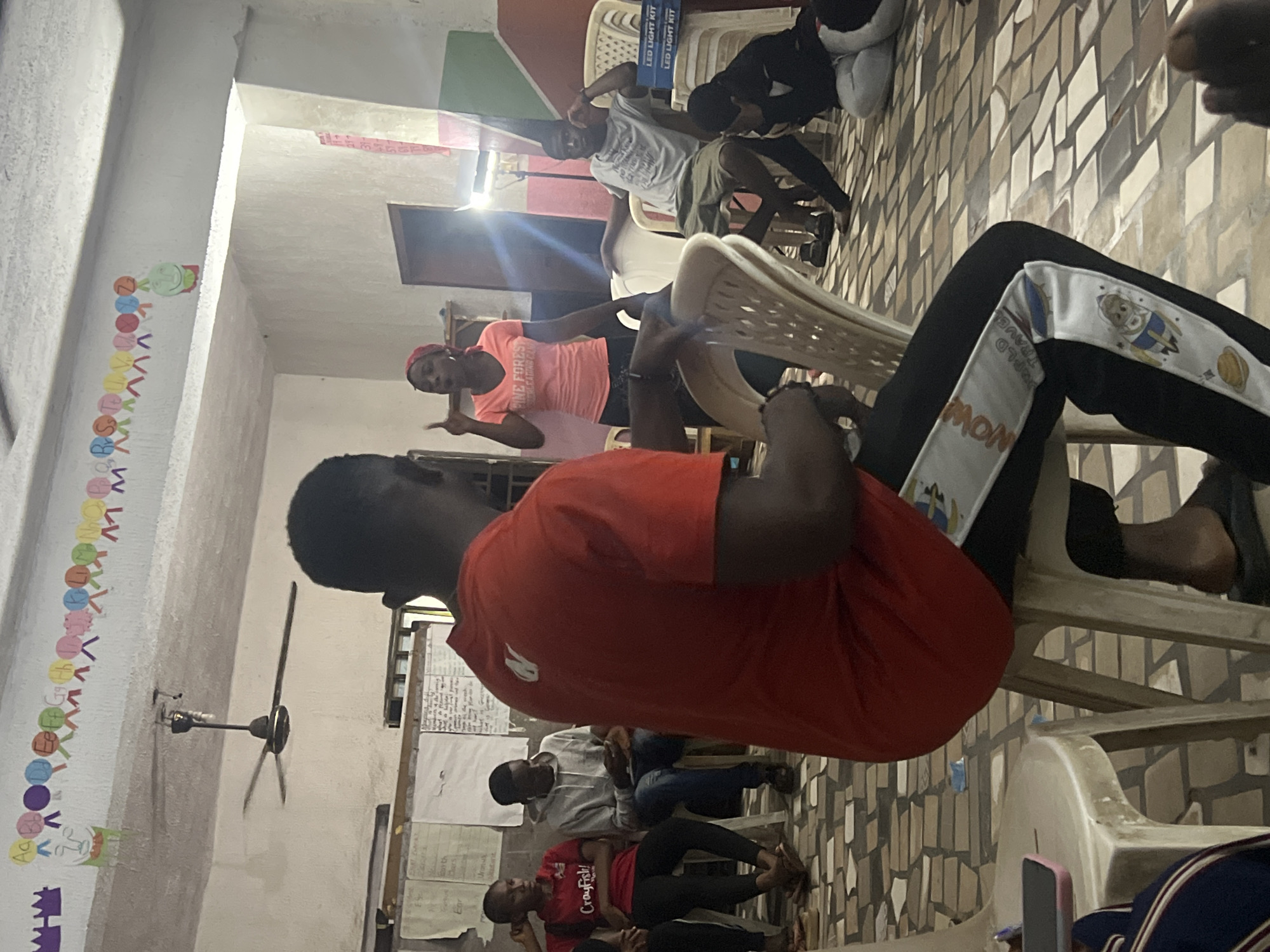 In Nkpor, Nigeria, a group of young people are gathered indoors, likely in a classroom or community center. The room has a tiled floor and is decorated with alphabet charts and colorful drawings on the walls. A ceiling fan hangs from the ceiling, and there's a bright spotlight illuminating part of the space.
In the foreground, a young man in a red t-shirt is seated, facing away from the camera. He's wearing black pants with a white stripe featuring cartoon images of astronauts and planets.
To his left, a young woman in a red t-shirt with the text "Crayfish!" is seated, looking towards the woman speaking. Next to her, a young man in a grey hoodie is seated, looking at his phone.
In the center, a young woman with a red headscarf is standing and speaking, gesturing with her hand. She's wearing a pink t-shirt with the text "Pine Forest."
To her right, a young man in a grey t-shirt and shorts is seated, resting his head on his hand. In the background, several white plastic chairs are stacked. On one of these chairs, two boxes labeled "LED LIGHT KIT" are visible. Further back, a young person in dark clothing is kneeling. The overall atmosphere suggests a meeting, presentation, or informal gathering.