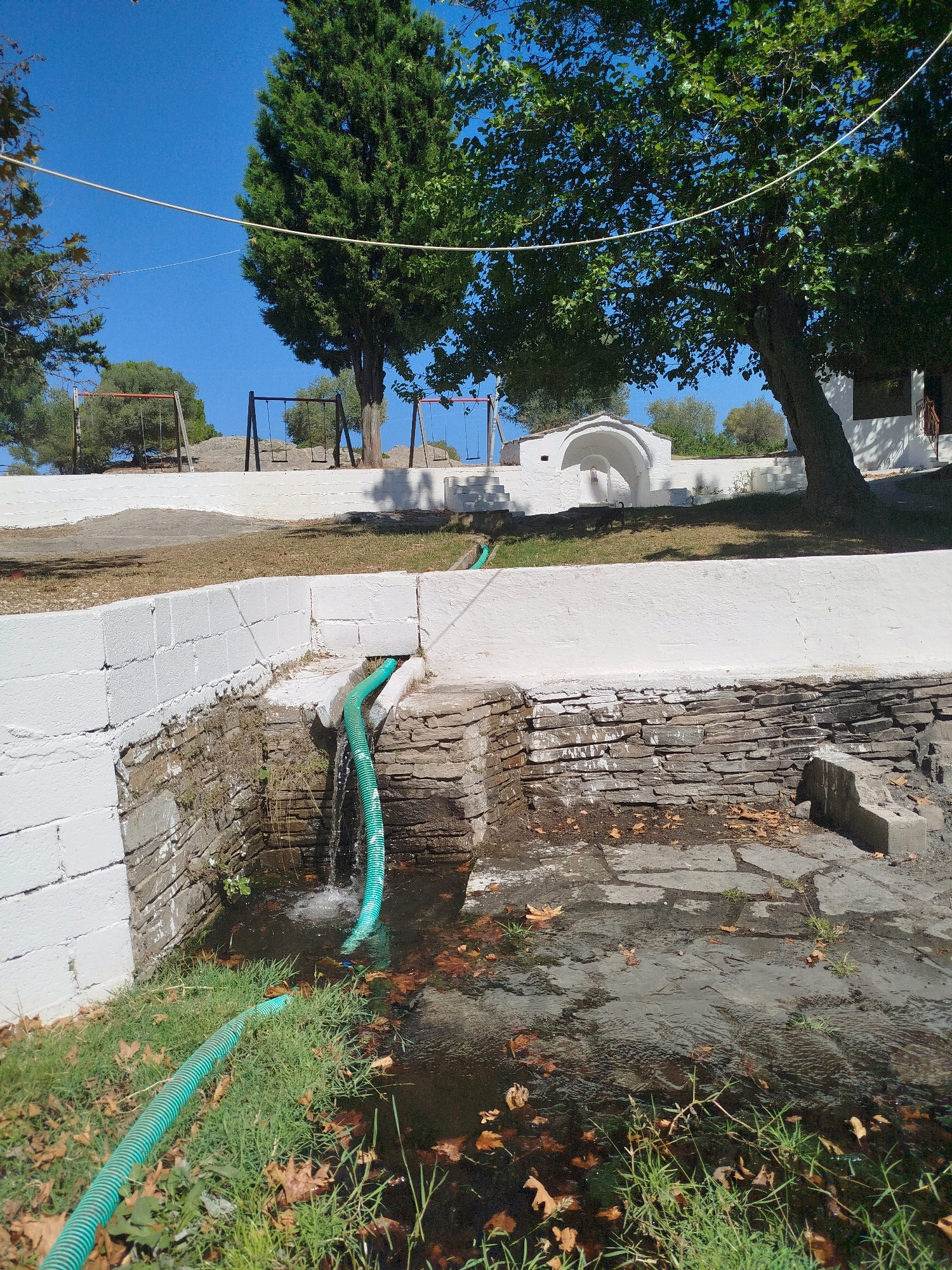 The image depicts an outdoor scene with a focus on a water feature. In the foreground, a green corrugated hose lies on lush green grass scattered with fallen brown leaves. The hose runs towards a collection of rough-hewn stones, forming a wall or basin. Water is visibly flowing from another green hose into a dark pool of water within this stone structure. The pool contains more fallen leaves and some sparse green vegetation.
Behind the water feature, a white-painted, block-constructed wall extends horizontally across the frame. Beyond this wall, a grassy area is visible, leading up to a row of swings. There are at least three sets of swings with red and black frames, set against a backdrop of trees and a clear blue sky. Further in the distance, a small white arched structure, possibly a fountain or shrine, is situated on a raised platform. A large tree with dense green foliage dominates the right side of the image, casting shadows on the white structure. The overall impression is of a public park or recreational area.
This scene is located in the Sithonia Municipal Unit, Greece.