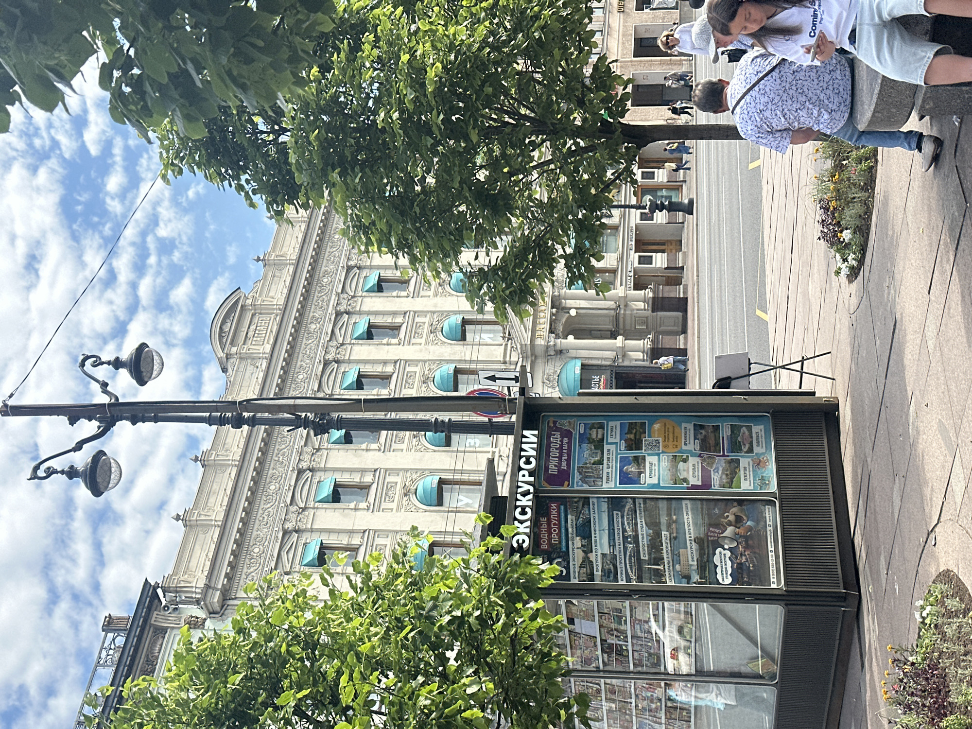 The image shows a street scene in front of a classical building. A kiosk offering excursions is visible on the left, displaying various tours and destinations. The building behind the kiosk has a light-colored facade with ornate architectural details and aqua-colored awnings over the windows. A lamppost with two globes stands prominently in the foreground. Green trees with lush foliage are scattered throughout the scene, some framing the view of the building. In the middle ground, a street with a few people walking and cars can be seen. In the foreground, a group of people are sitting on a stone bench. A woman in a white top and light blue shorts is looking at her phone, and a man in a blue and white patterned shirt is seated next to her.