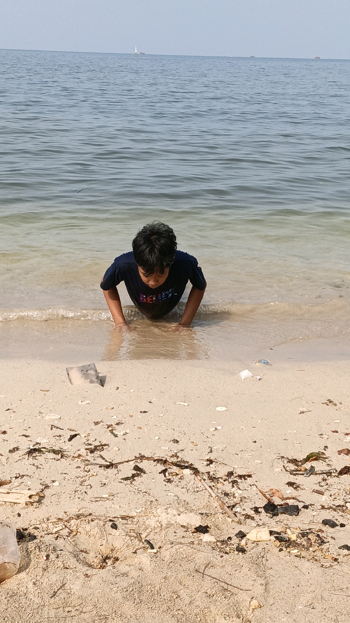 A young boy with dark hair is in a push-up position in the shallow water at the edge of a sandy beach, with his hands on the seabed and his torso submerged. He is wearing a dark blue t-shirt with a colorful graphic design. The water is calm and clear, with small waves lapping at the shore. The beach is covered in sand, with some scattered debris, including seaweed, shells, and what appear to be pieces of plastic and wood. In the distance, the sea stretches to the horizon under a pale sky, with a few small boats visible. The overall scene depicts a boy playing at the water's edge on a bright day.