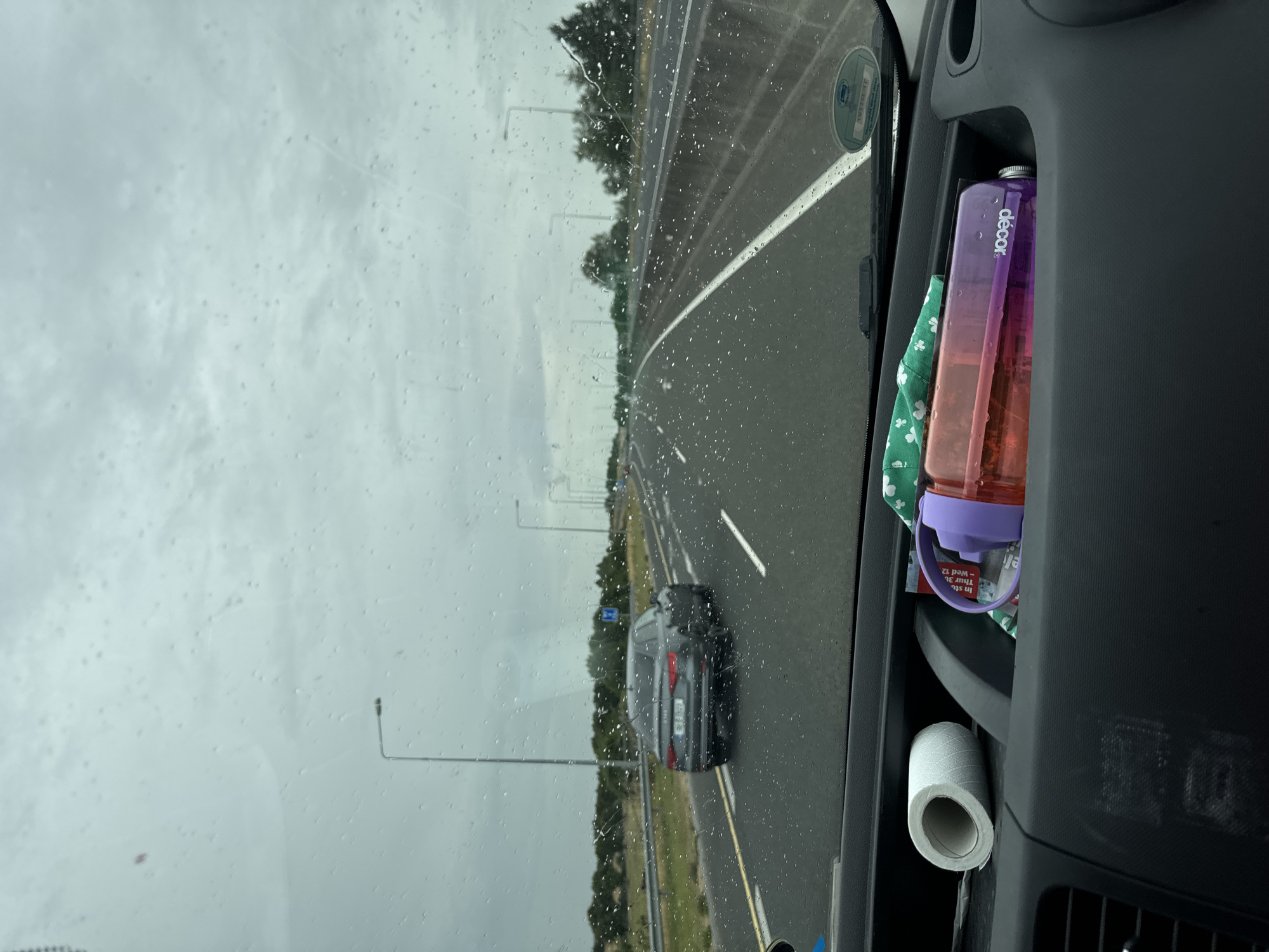 The image depicts a view from inside a vehicle looking forward through a rain-streaked windshield onto a highway. A dark-colored Audi sedan is visible driving in the right lane ahead. The highway has multiple lanes and is lined with trees and streetlights. In the foreground, inside the vehicle, a roll of toilet paper and a reusable water bottle with a purple lid and orange body are visible in a compartment. The sky is overcast and grey. The location is identified as Dunbell, Ireland.