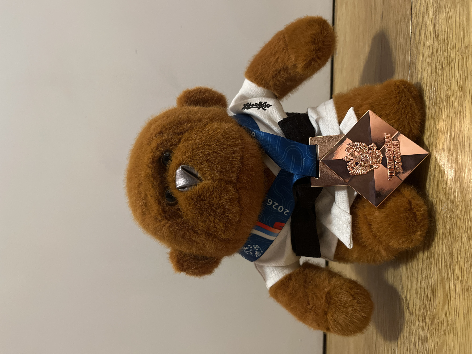 A brown teddy bear wearing a white shirt, black vest, blue lanyard with "2026" printed on it, and a bronze-colored medal with an emblem of Russia rests on a surface. The location is not discernible from the image.