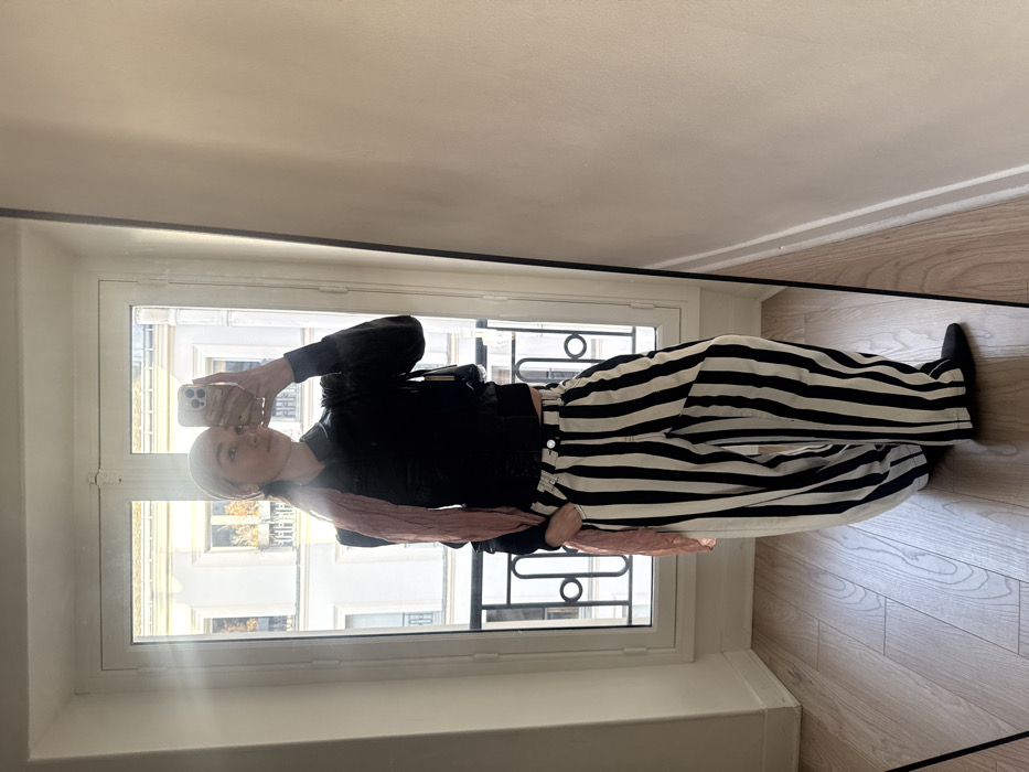 A female subject is captured in a mirror selfie, standing indoors with a smartphone raised to photograph her reflection. She wears a dark long-sleeved top, wide-leg trousers with vertical black and white stripes, and black flat shoes. A light pink scarf is draped over her head and left shoulder. A small black handbag hangs on her left side, and her left hand is positioned in her trouser pocket. The indoor setting features light-colored walls and a light-toned wooden floor. Behind the subject, a large, white-framed window provides a view of typical Parisian Haussmannian architecture, including light-colored stone buildings, multi-story facades, and a black wrought-iron balcony railing immediately outside the window. Distant buildings and trees with visible autumnal foliage complete the background, indicating a street scene in Paris, France, under natural daylight.