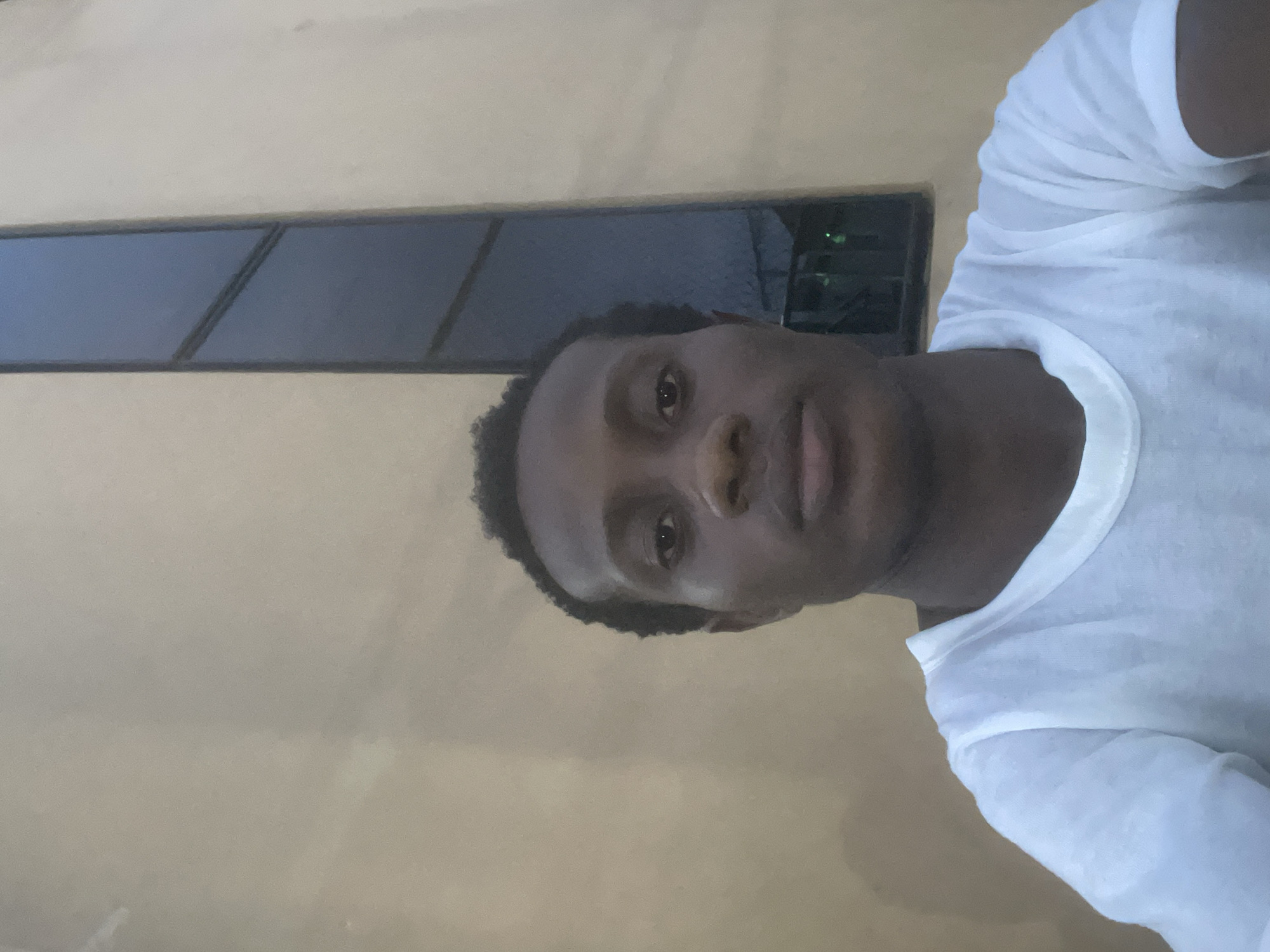 The image is a selfie of a young Black man wearing a white t-shirt. He is standing in front of a building with a light brown, textured wall. To the right of the man, a tall, narrow window with dark glass is visible, set into the building's facade. The lighting suggests it is either early morning or late afternoon. The setting appears to be an outdoor urban or semi-urban environment in Ihiagwa, Nigeria. There are no other individuals or significant objects in the frame.