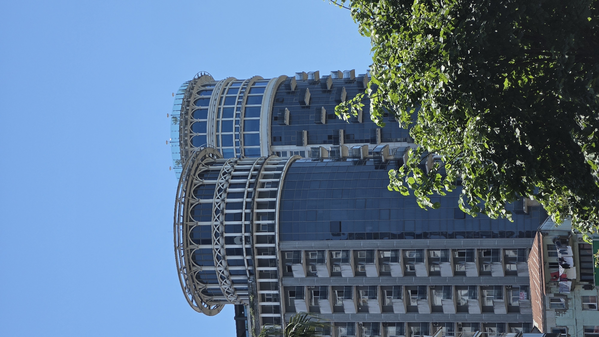 The image shows two distinctive, modern high-rise buildings in Batumi, Georgia. Both buildings have rounded, cylindrical structures with multiple floors. The building in the foreground on the left has a predominantly grey exterior with numerous balconies and dark blue glass windows. Its upper section features an ornate, circular white decorative element with arched windows. The building behind it on the right shares a similar rounded design and a blue glass facade, but its upper portion is adorned with a modern, open-air terrace featuring a glass railing and some greenery. In the lower part of the image, there are glimpses of lush green trees and a smaller, older building with a rusted metal roof and laundry hanging on a balcony, providing a contrast to the modern architecture. The sky is a clear, bright blue, indicating a sunny day.