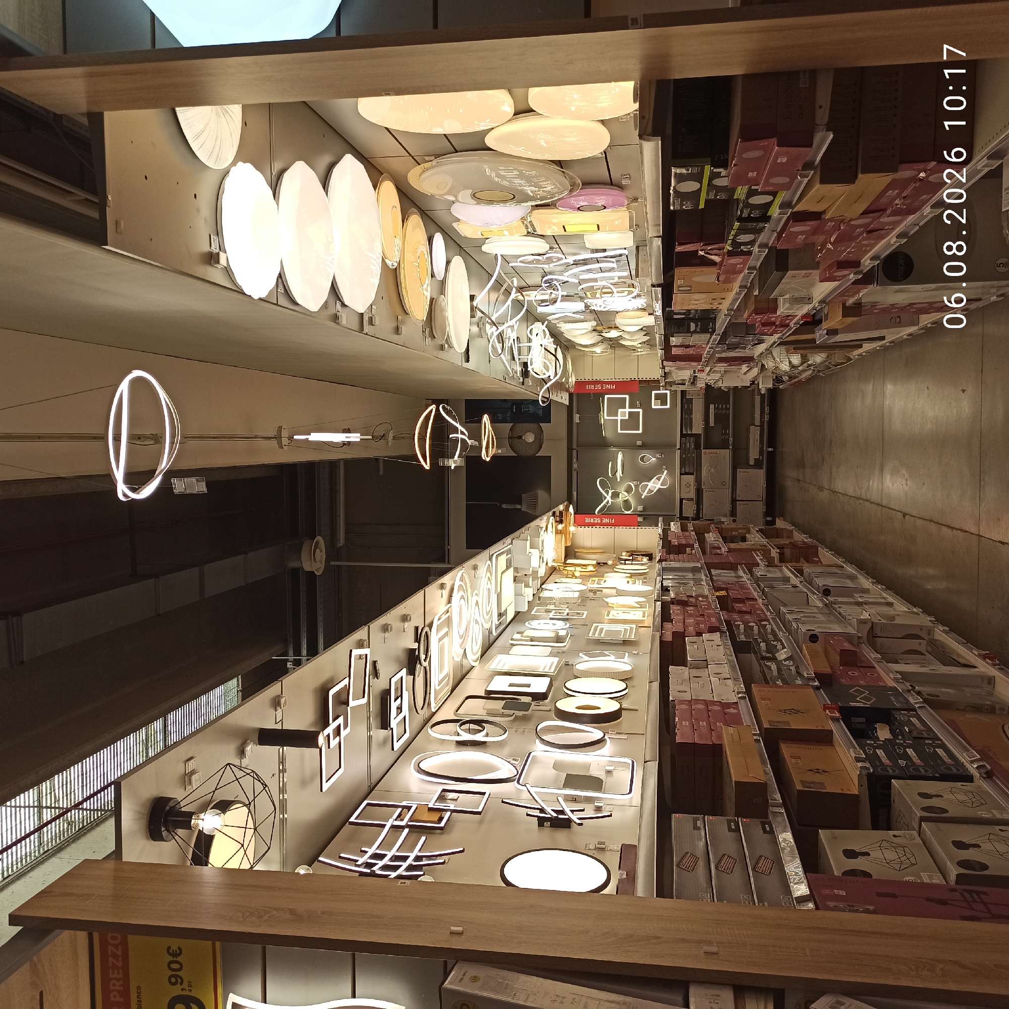 The image shows an aisle in a retail store, likely a home improvement or lighting store, with shelves on either side displaying various lighting fixtures. The aisle is long and appears to be in a commercial setting. On the left, there are displays of ceiling lights and other decorative lighting fixtures. Some boxes are stacked on lower shelves. On the right, there are more shelves filled with various lighting fixtures, including modern, geometric designs and more traditional-looking ceiling lights. In the background, the aisle continues, with more shelving and lighting displays visible. At the end of the aisle, there appear to be large display boards or screens. The lighting in the store is a mix of overhead fluorescent lights and the light emitted from the displayed fixtures themselves. The floor is concrete. There are no people visible in the image. The timestamp "06.08.2026 10:17" is overlaid on the bottom right corner. The image is a wide-angle shot, capturing a significant portion of the aisle.