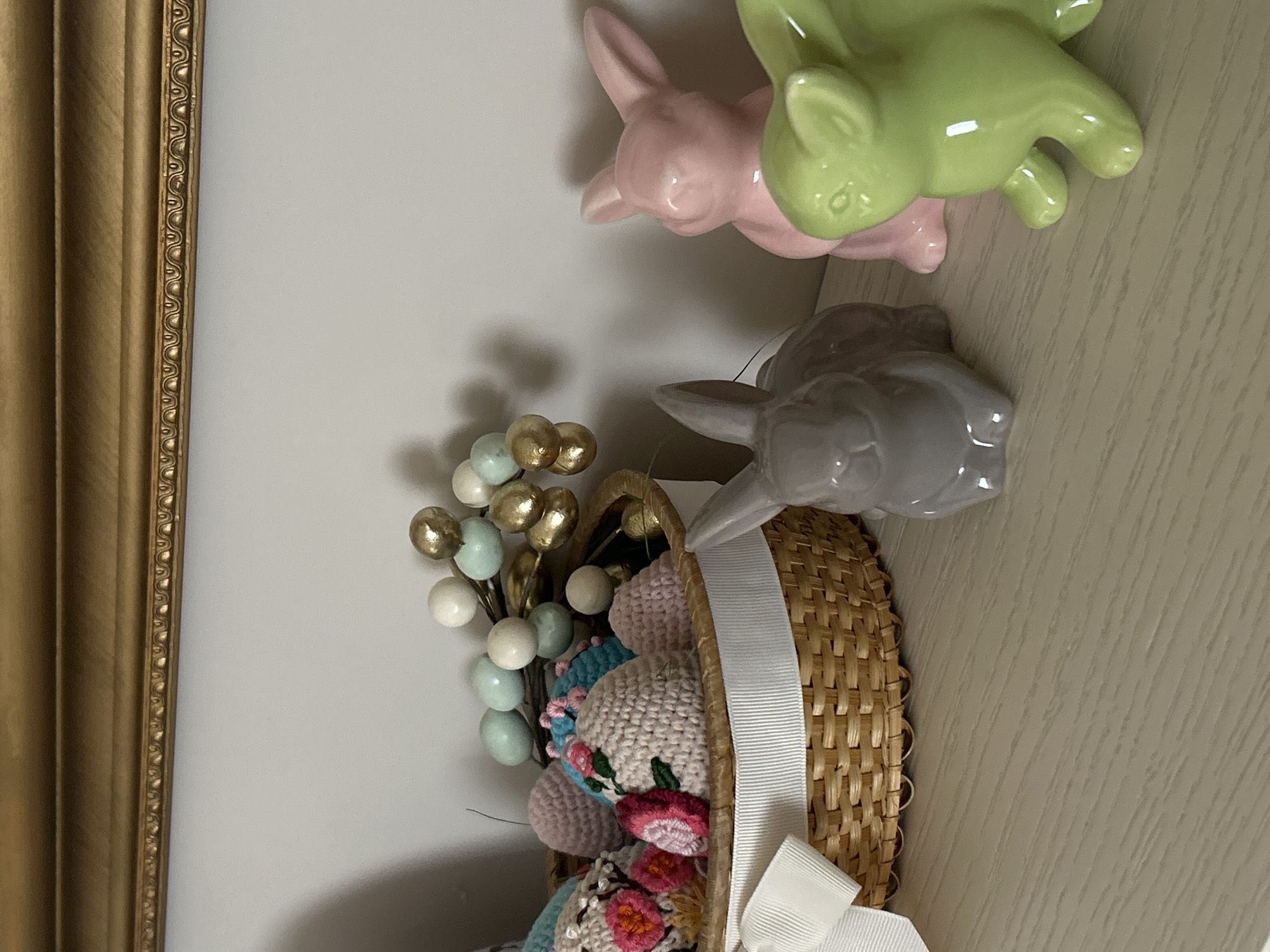 The image shows a close-up of an Easter-themed decorative arrangement. There is a wicker basket filled with crocheted Easter eggs adorned with colorful floral patterns. Emerging from the basket are decorative twigs with small, round embellishments in colors like gold, light blue, and white. Three ceramic rabbit figurines are positioned near the basket's rim, in pink, green, and gray. A white ribbon is tied around the basket. A section of a gold-colored ornate frame can be seen on the left side of the image. There is no information indicating the location being Plovdiv, Bulgaria.
