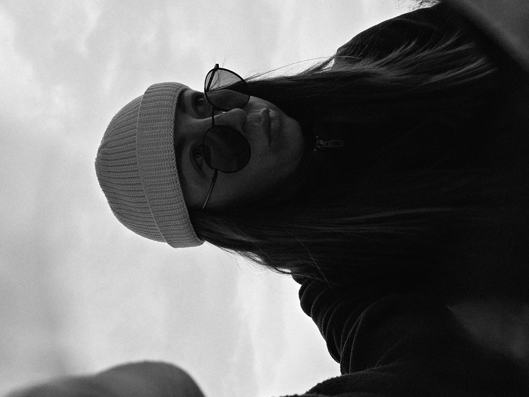 A low-angle, black and white photo shows a person wearing a beanie and sunglasses. The person has long hair and is wearing a jacket with a visible zipper. The background is a cloudy sky. The location is unknown.