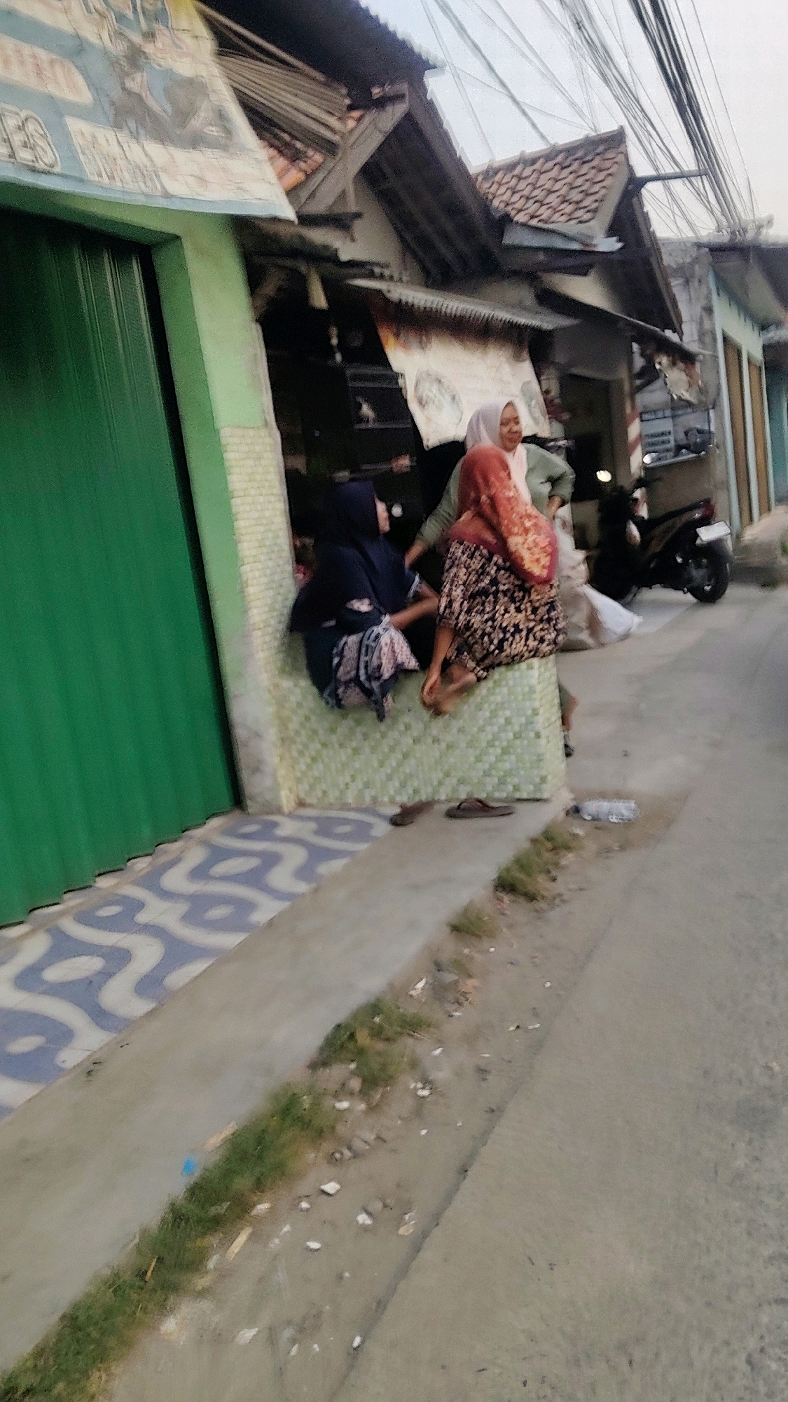 In the city of Mekarmukti, Indonesia, three women are seated on a low, tiled wall outside a shop. The shop's facade features a vibrant green door and a portion of a banner with a graphic. The women are dressed in traditional Islamic attire, with two wearing hijabs. One woman is seated on the far left, wearing a dark hijab and a patterned skirt. The woman in the middle wears a light green top and a patterned red and orange hijab. The woman on the right is seated with her back to the viewer, wearing a dark and light patterned skirt and a red hijab. To the right of the shop, a motorcycle is parked. The scene is situated on a paved street with a concrete sidewalk. The environment appears to be a commercial area, with a sense of everyday life unfolding.