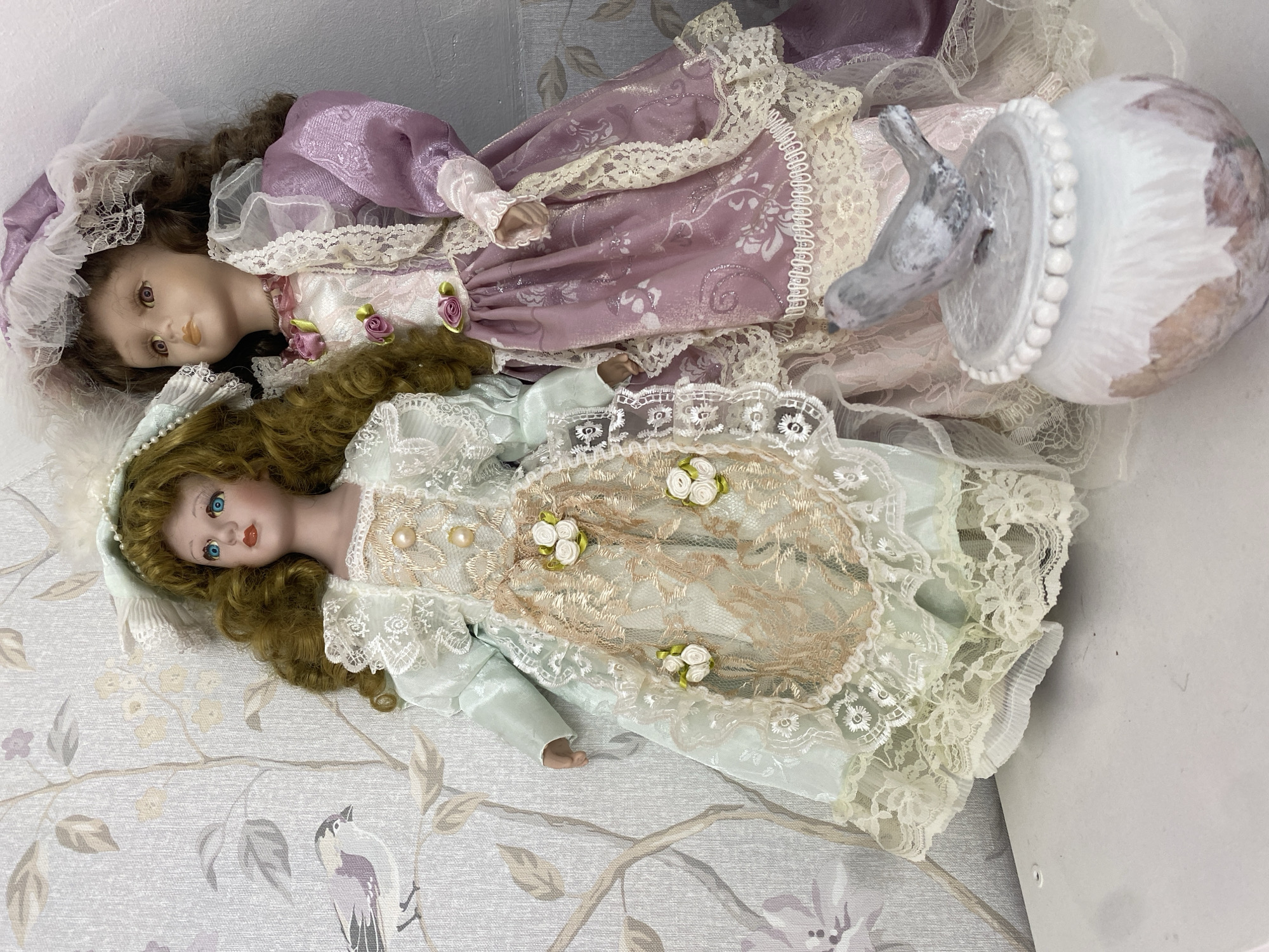 The image displays two porcelain dolls lying on a patterned surface. The doll positioned higher has dark brown, curly hair and purple eyes. She wears an elaborate purple dress with a shimmering pattern, extensive white lace trim, and small pink rose appliques, topped with a matching purple bonnet adorned with lace. The second doll, positioned below, has lighter brown, wavy hair and bright blue eyes. She is dressed in a light green gown with gold lace overlay, detailed with white pearls and small white ribbon flowers, and features intricate lace sleeves and trim.
The dolls are resting on a light-colored surface that appears to be fabric or wallpaper, patterned with delicate grey branches, leaves, and stylized birds in muted purple and grey tones. To the right, a decorative element shaped like a grey and white bird is perched on a white, textured spherical base with a beaded edge. The scene is an indoor setting, likely a display area for collectible dolls. No specific activity is occurring; the dolls are posed for viewing. There is no visible text in the image.