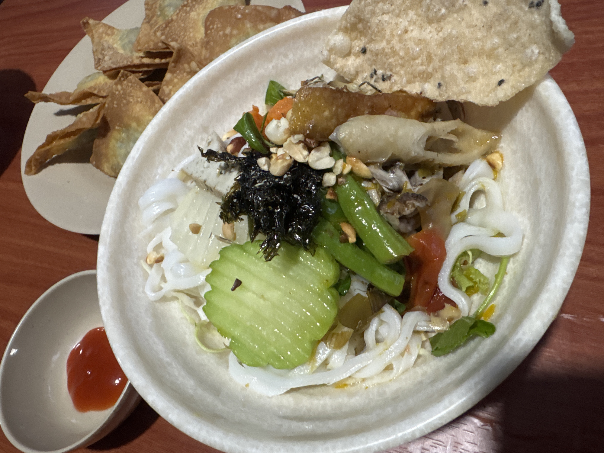 The image shows a plate of Vietnamese Mì Quảng (turmeric noodles) in a white bowl, garnished with green beans, cucumbers, peanuts, and a crispy cracker. Beside it is a smaller bowl with a red dipping sauce, and a plate of fried wontons. The setting appears to be a restaurant in Đà Nẵng, Vietnam. The background consists of a dark red wooden table.